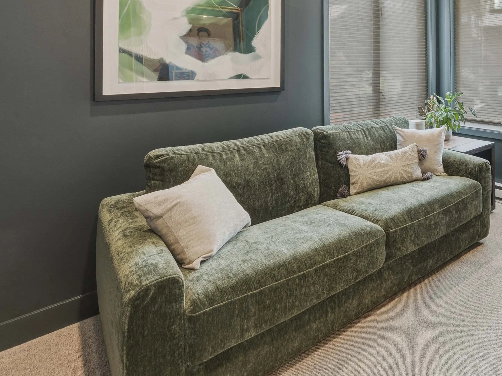 A green upholstered sofa with three decorative pillows in a room with gray walls, large windows with blinds, a side table with a plant, and a framed picture hanging on the wall.