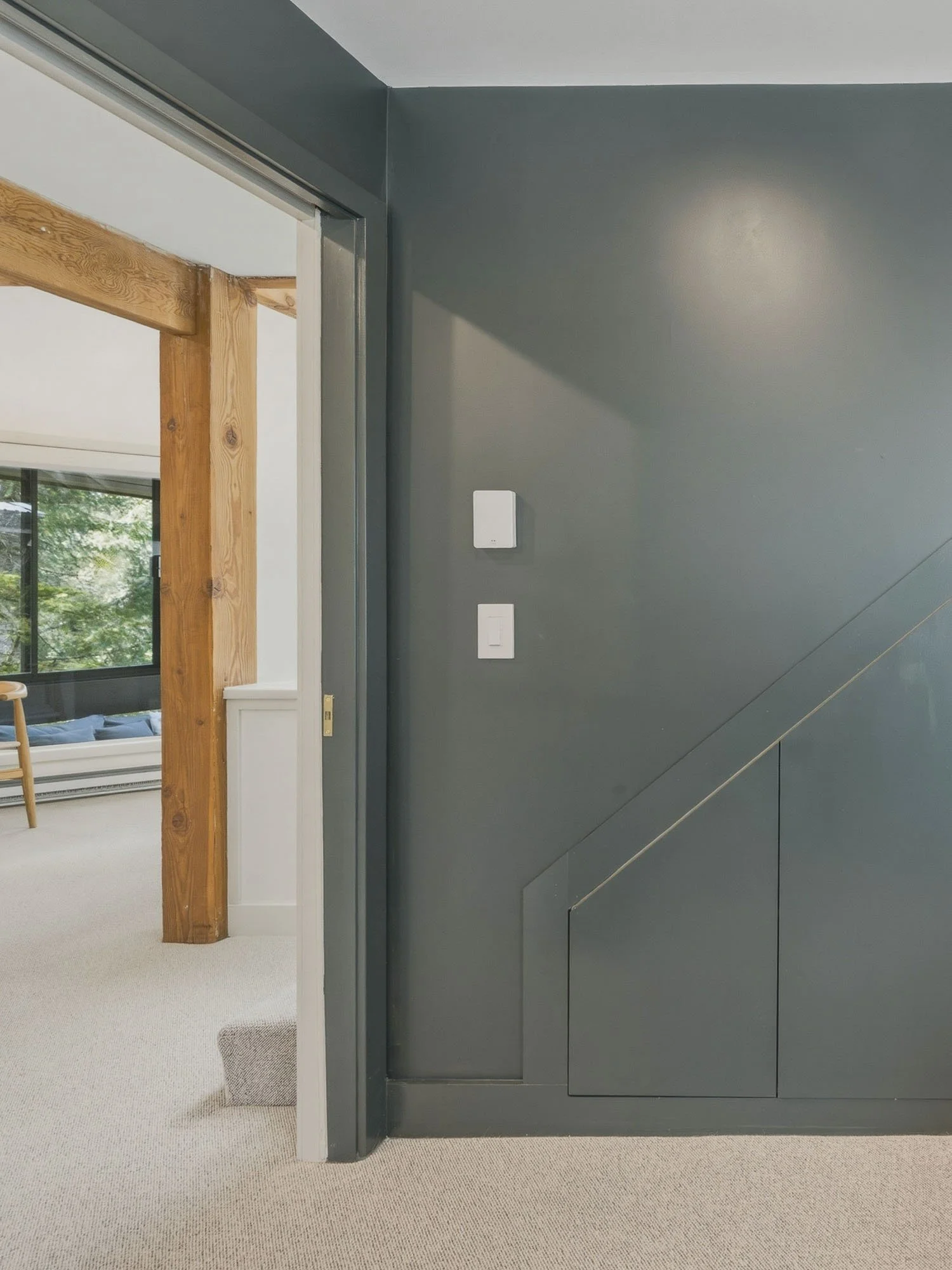 Interior view with a gray wall and a corner switchplate, adjacent to a doorway revealing part of a room with wooden beams, a window, and a chair.
