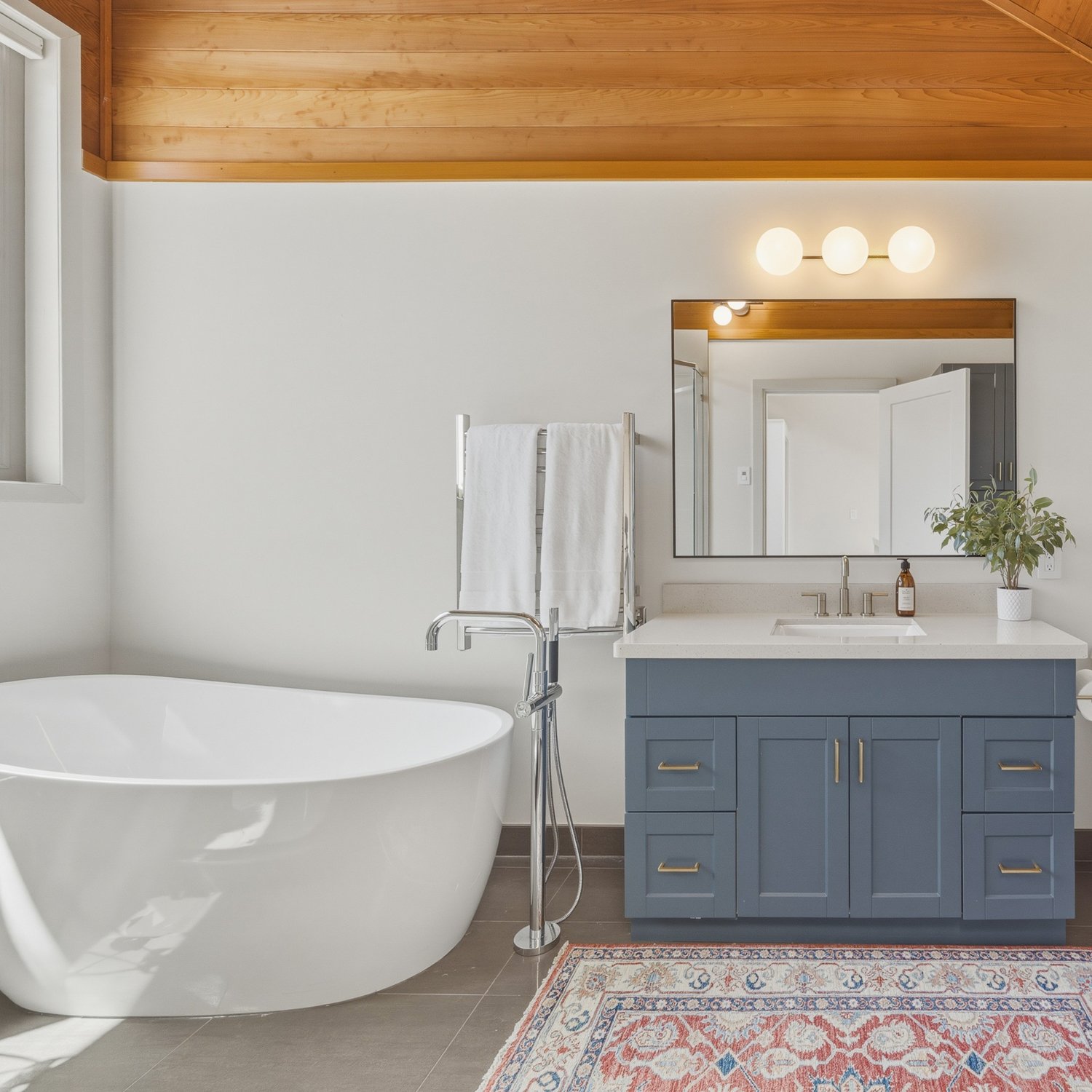 Modern bathroom with a white bathtub, a blue vanity with a white countertop, a large mirror, and a potted plant. There are white towels hanging on a rack and a rug on the floor. The ceiling has wood paneling, and there are three round lights above the mirror.
