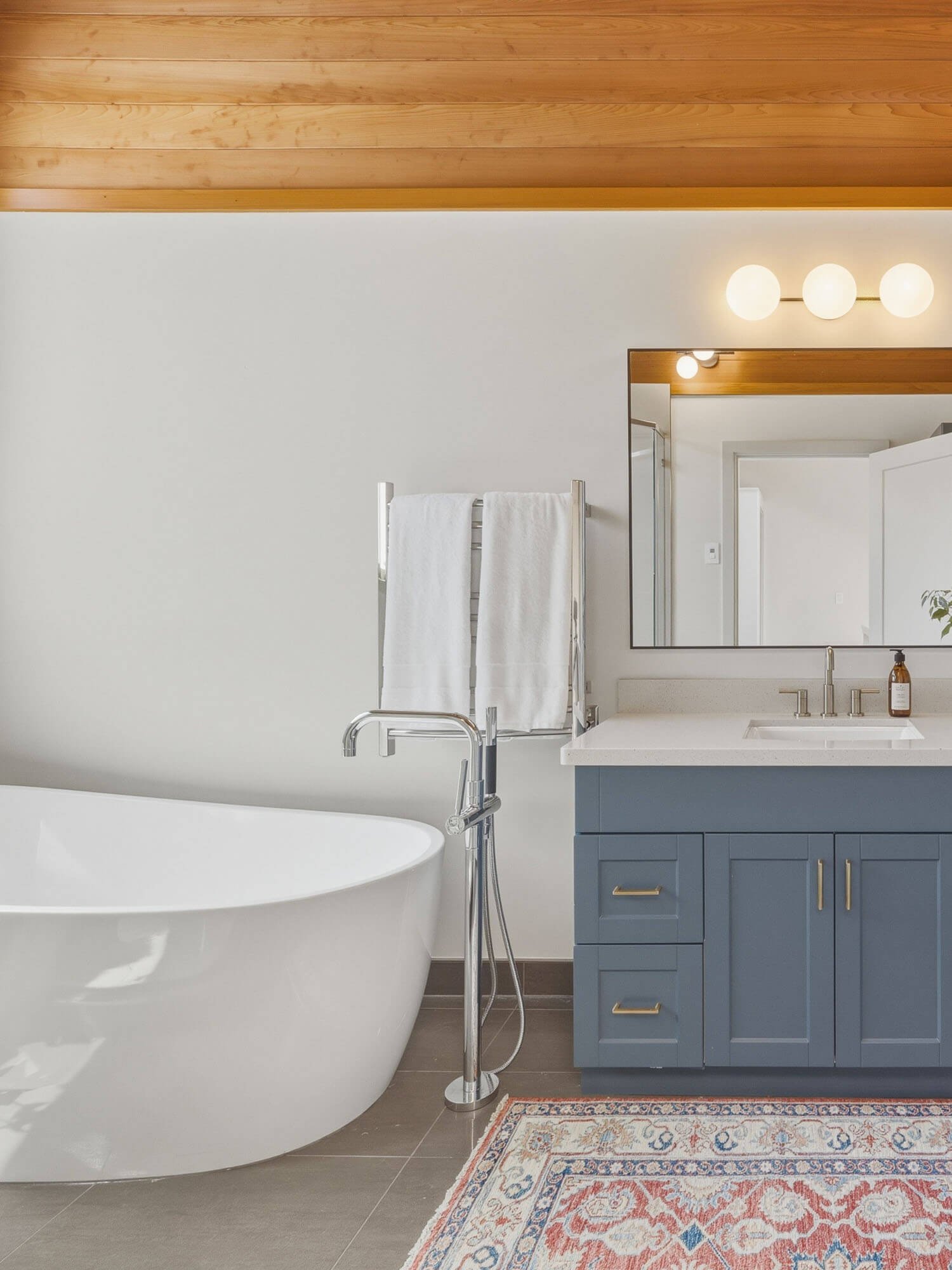 Modern 3-piece bathroom at 6117 Eagle Drive featuring a deep white bathtub, custom blue vanity cabinetry, programmable heated tile flooring, and a warm wood panel ceiling finish.