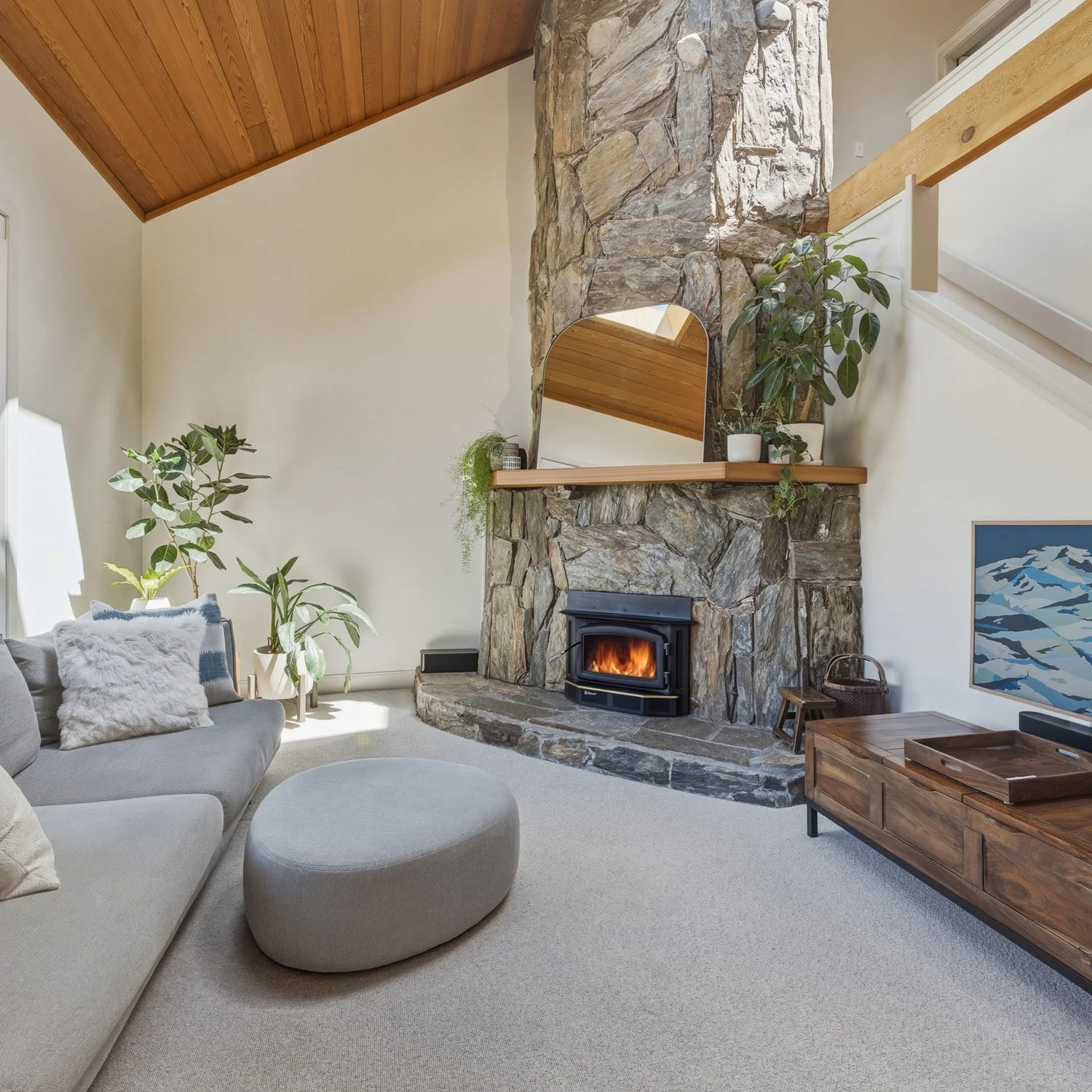 Living room with a gray sofa, a matching round ottoman, and decorative pillows. A stone fireplace with a wood-burning stove and a wooden mantel decorated with potted plants. Large windows, a forest painting, and a wooden coffee table are also visible.