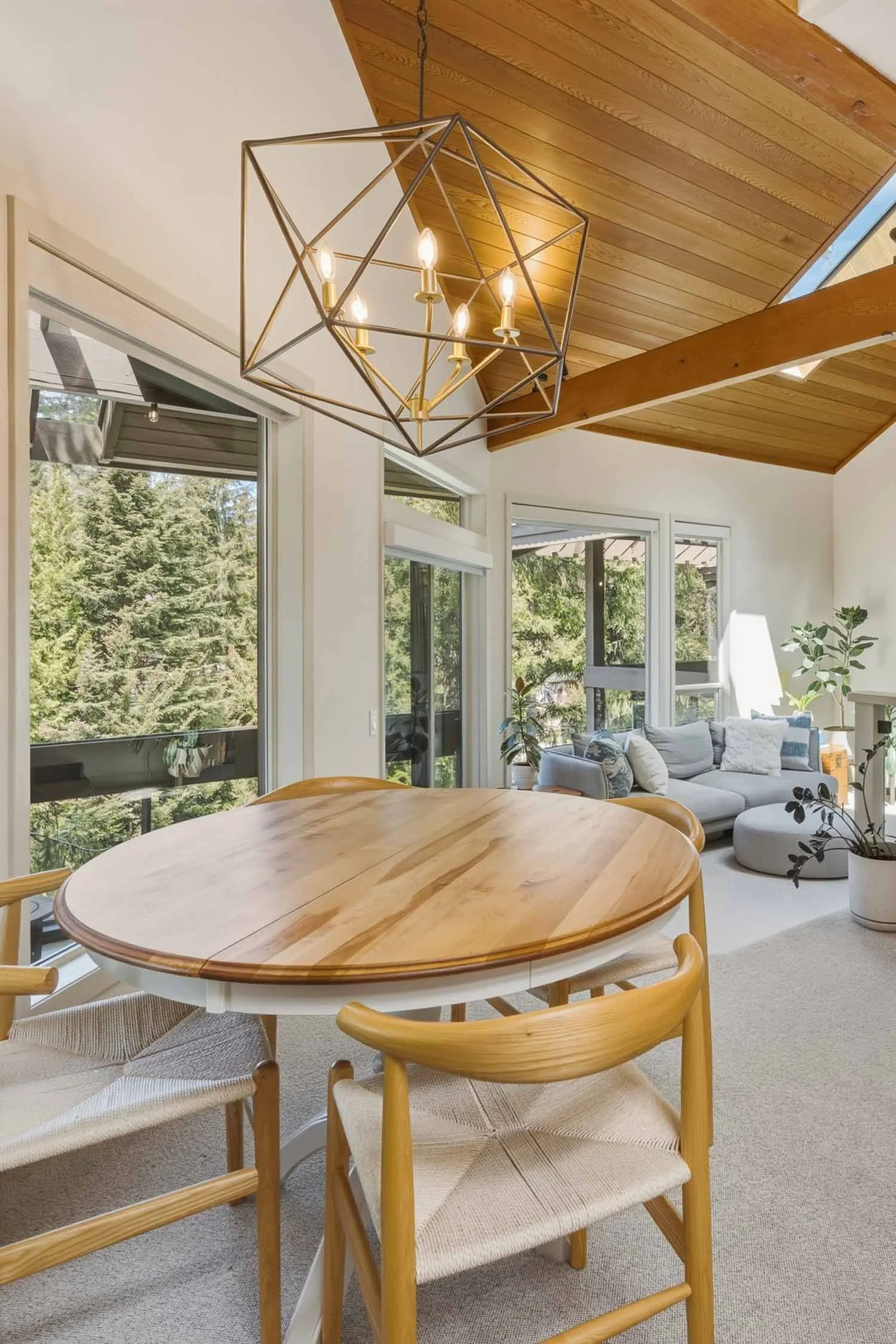 Intimate dining room space at Sunrise Whistler Cay Heights, perfect for entertaining, beneath a vaulted wood ceiling, custom lighting, and large windows framing the mature evergreen forest outside.