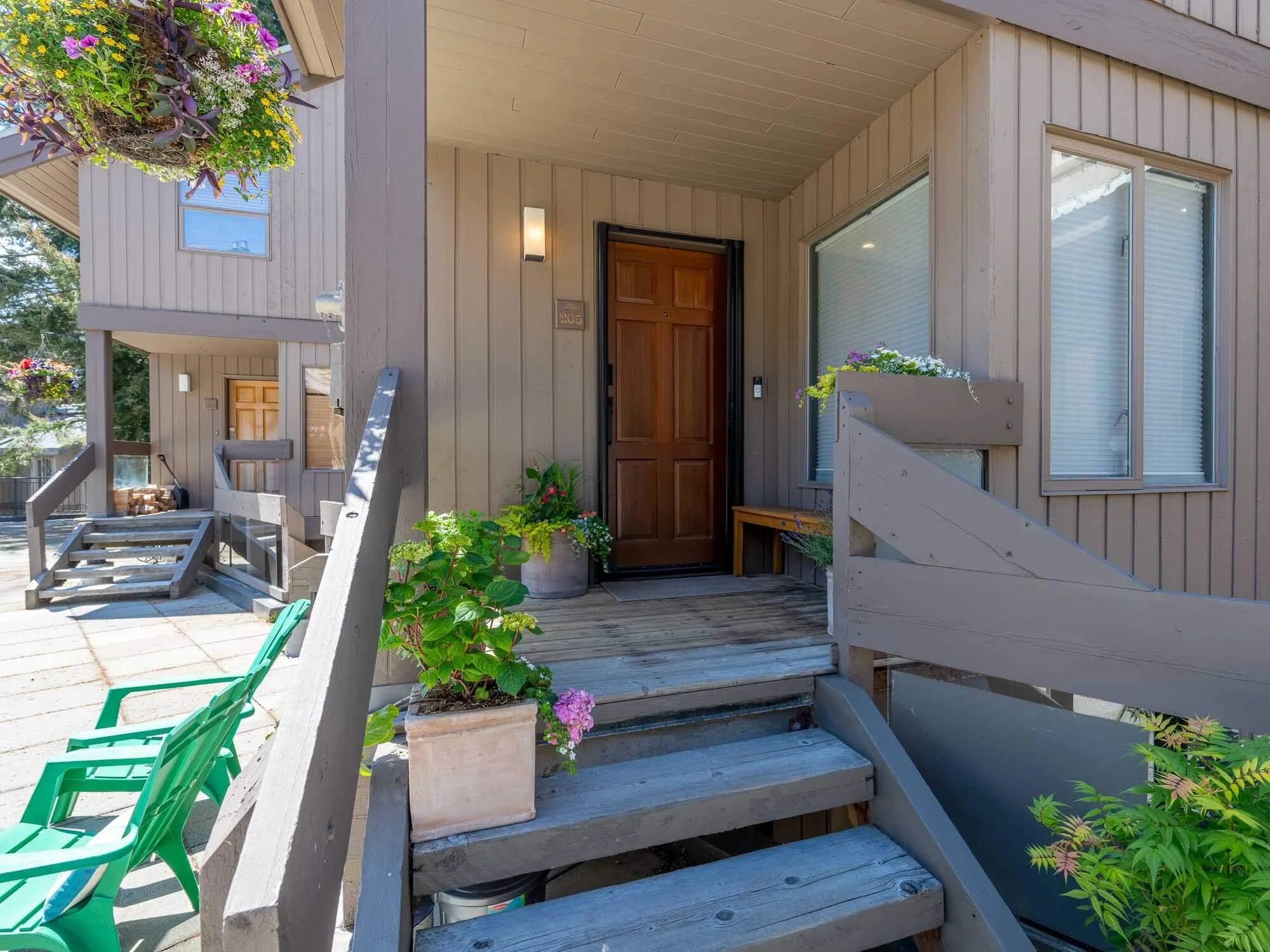 Front porch of a house with wooden stairs leading up to a green door, surrounded by potted plants and flowers, with a large window on the right side, and colorful hanging flower baskets on the upper left.