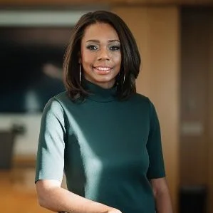 A woman with shoulder-length dark hair wearing a teal turtleneck sweater, smiling, in an indoor setting with wooden and modern decor.