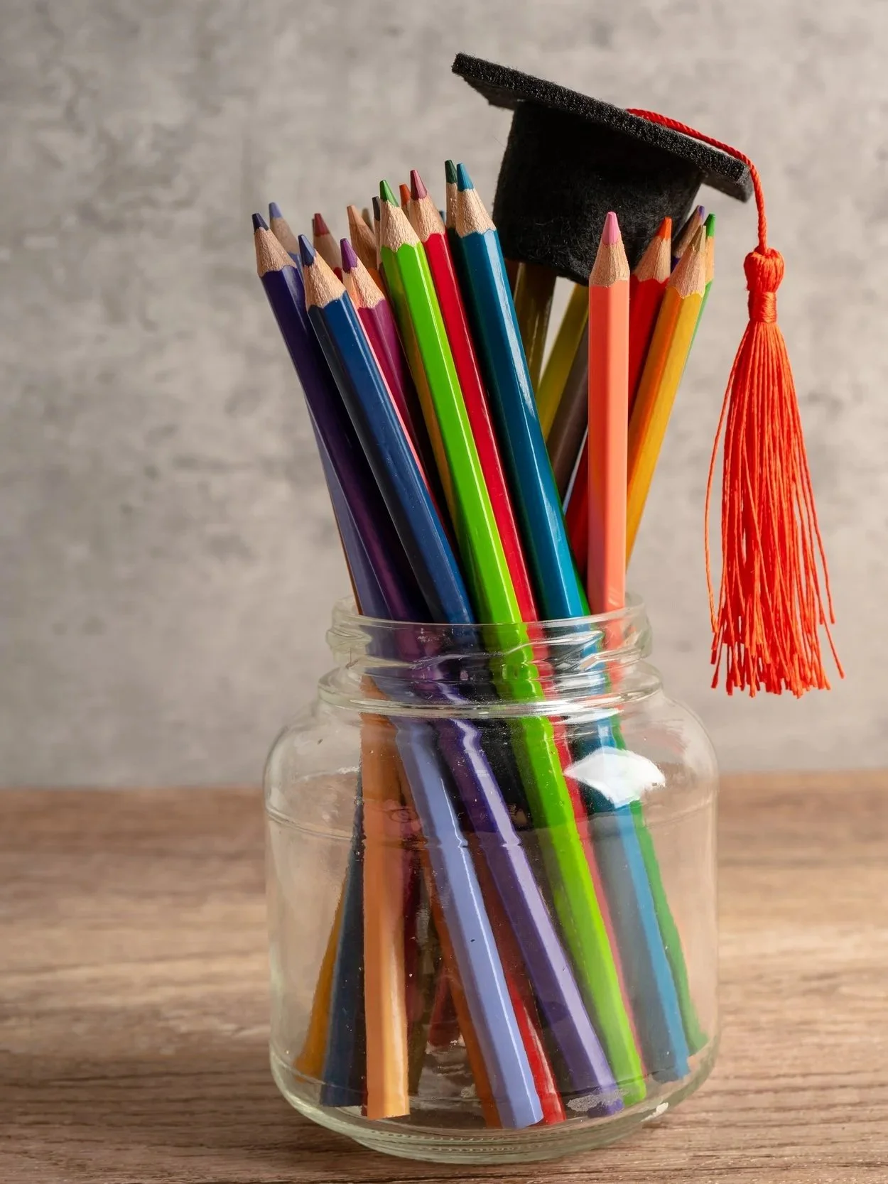 A glass jar holding colorful pencils, with a small black graduation cap with a red tassel on top of the pencils.