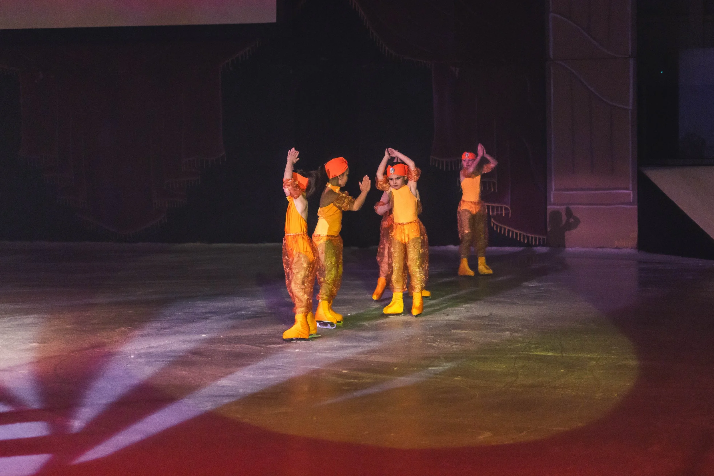 Four young girls wearing orange costumes and yellow ice skates are performing on an ice rink, with a dark stage background.