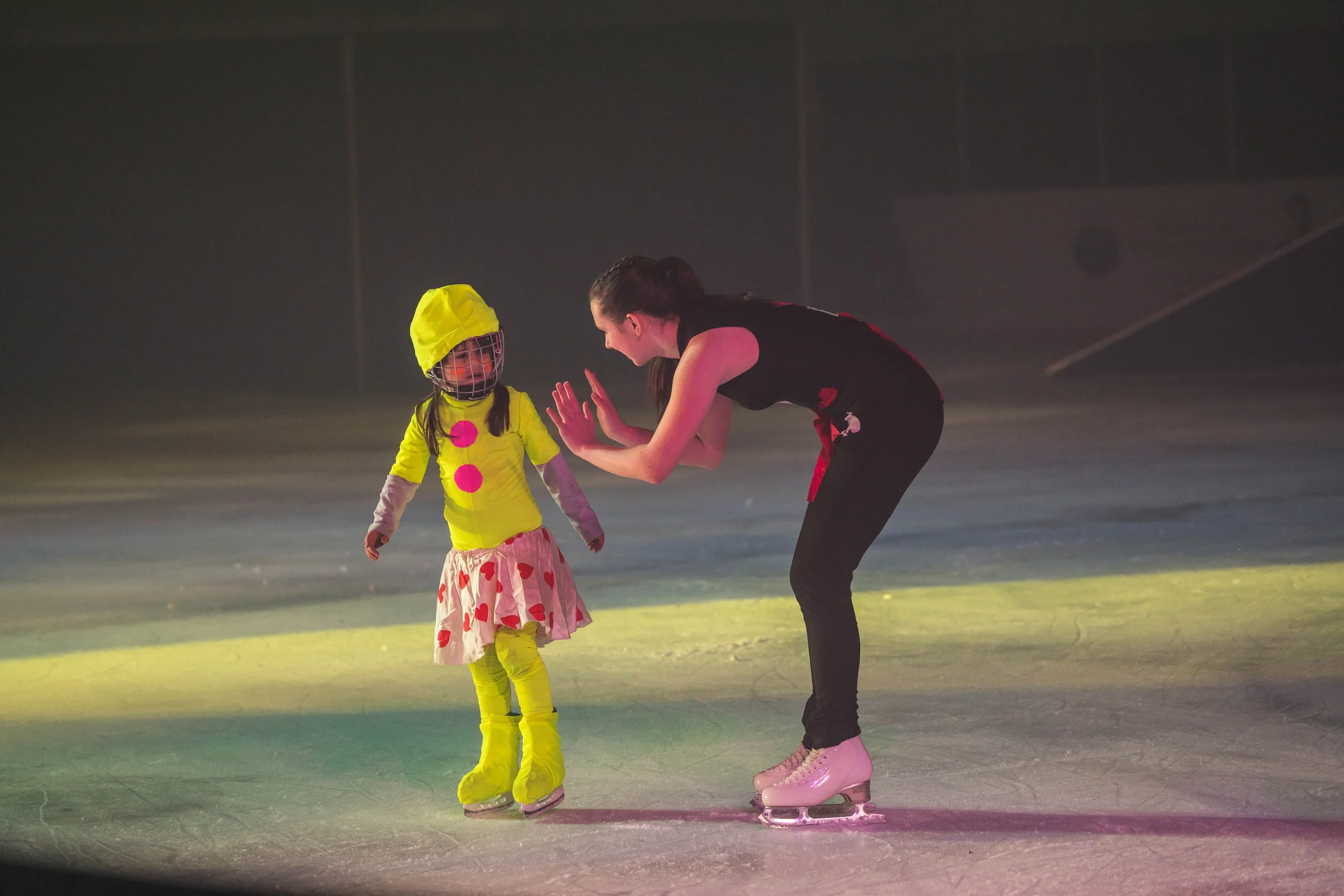 A woman and a young girl ice skating inside a skating rink. The girl is wearing a yellow outfit with pink dots and a yellow hood, and the woman is dressed in a black top and pants, both wearing ice skates, engaged in a training or coaching session.