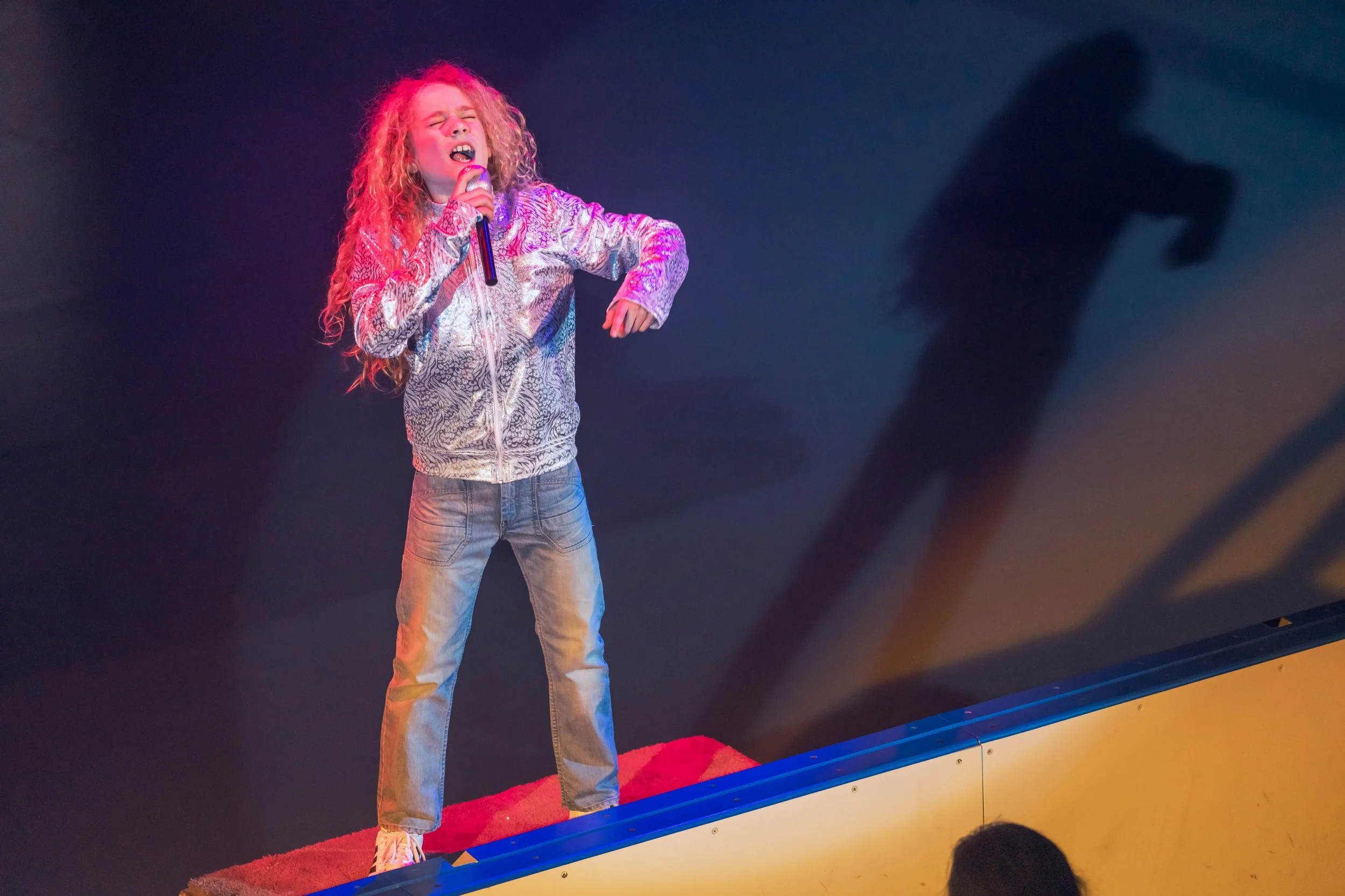 A young girl with curly hair singing passionately into a microphone on stage, wearing a patterned jacket and jeans, with colorful stage lighting.