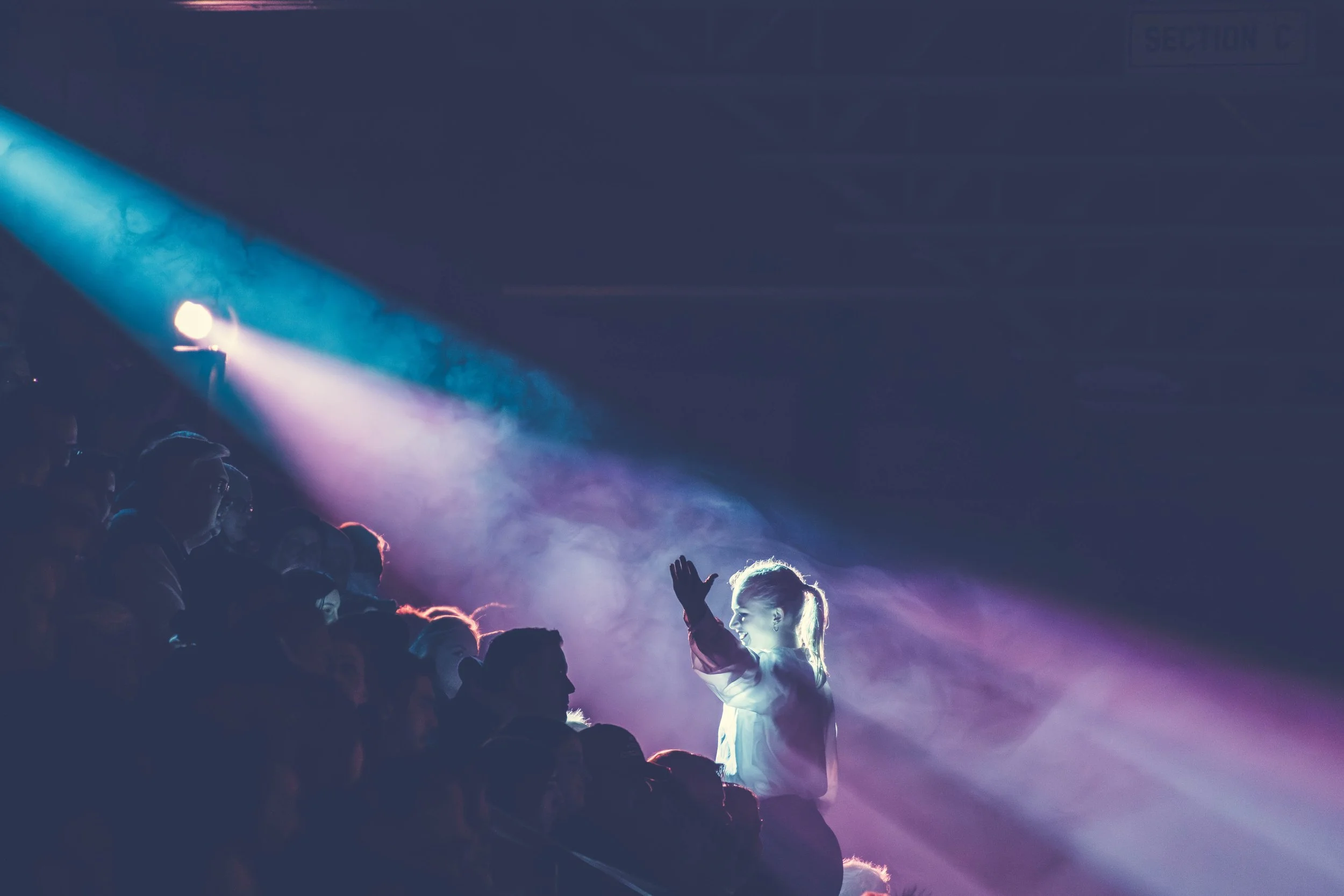 A crowd of people at a concert or event with a young girl on stage reaching out towards a colorful stage light.