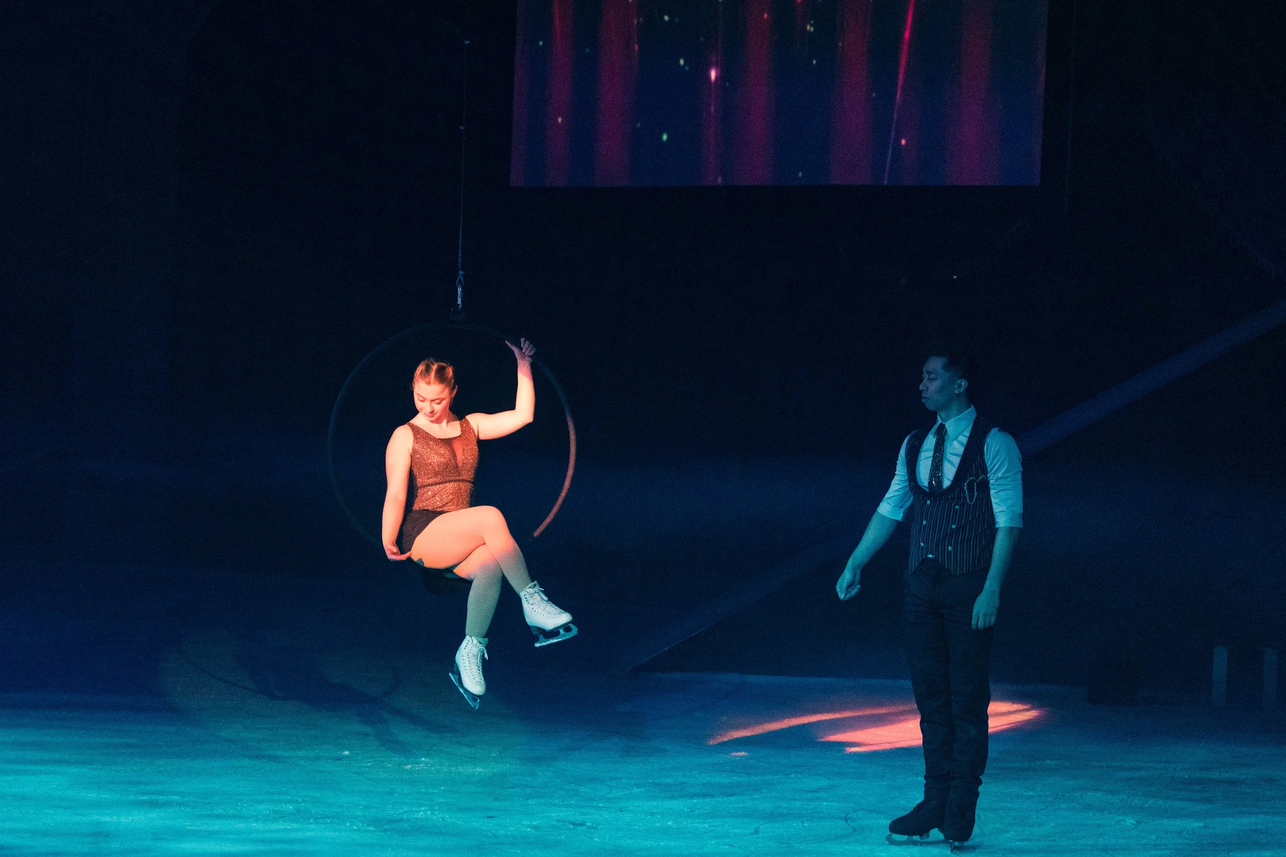 A female figure skater in a black and red outfit performing a jump on ice, with a male performer standing nearby with a striped vest and white shirt, in a dark, lit-up setting.