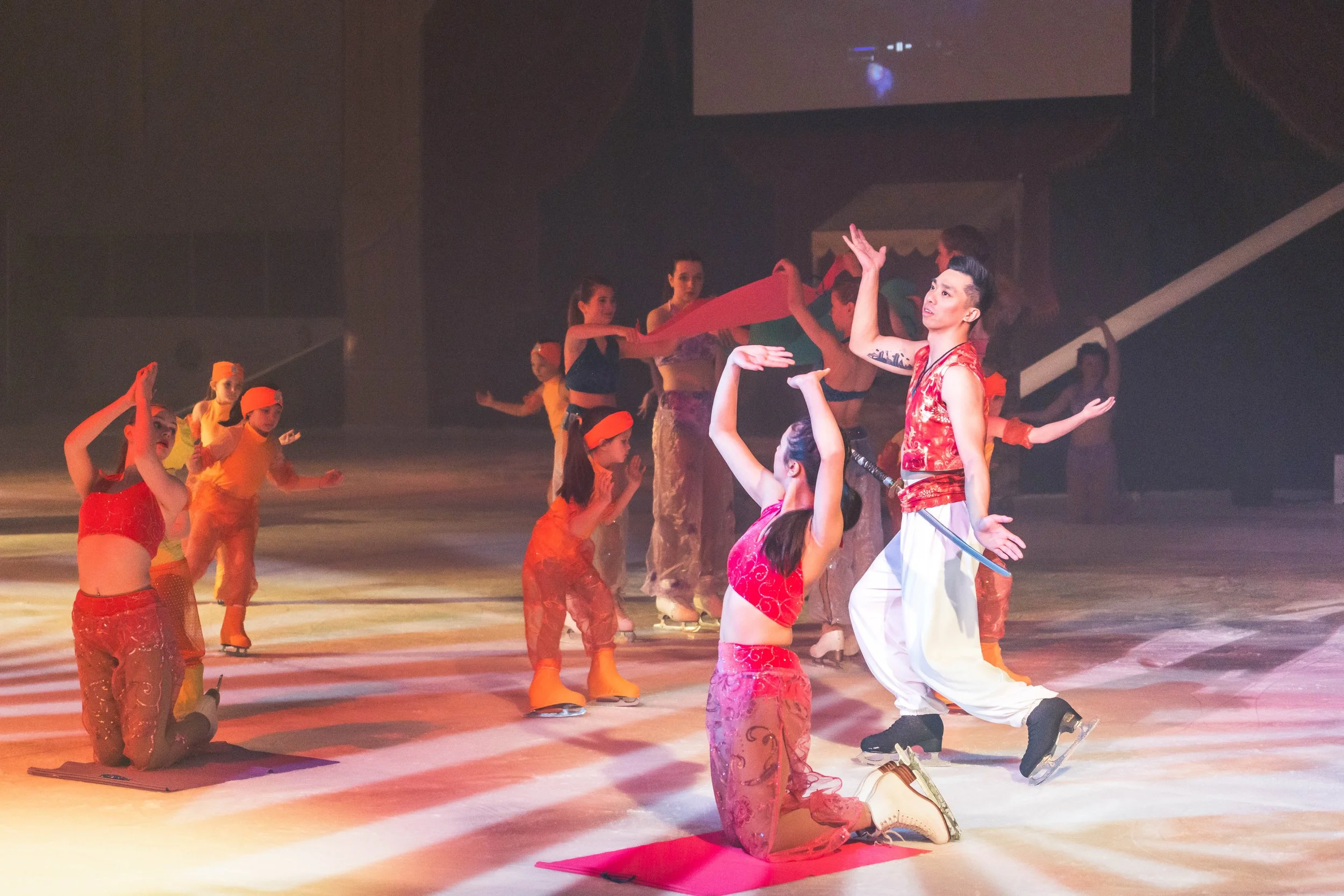 A group of performers dressed in colorful costumes practicing a routine on stage. Some are kneeling while others stand, with one performer in focus extending their arm upwards. The stage has warm lighting, and a large screen in the background display