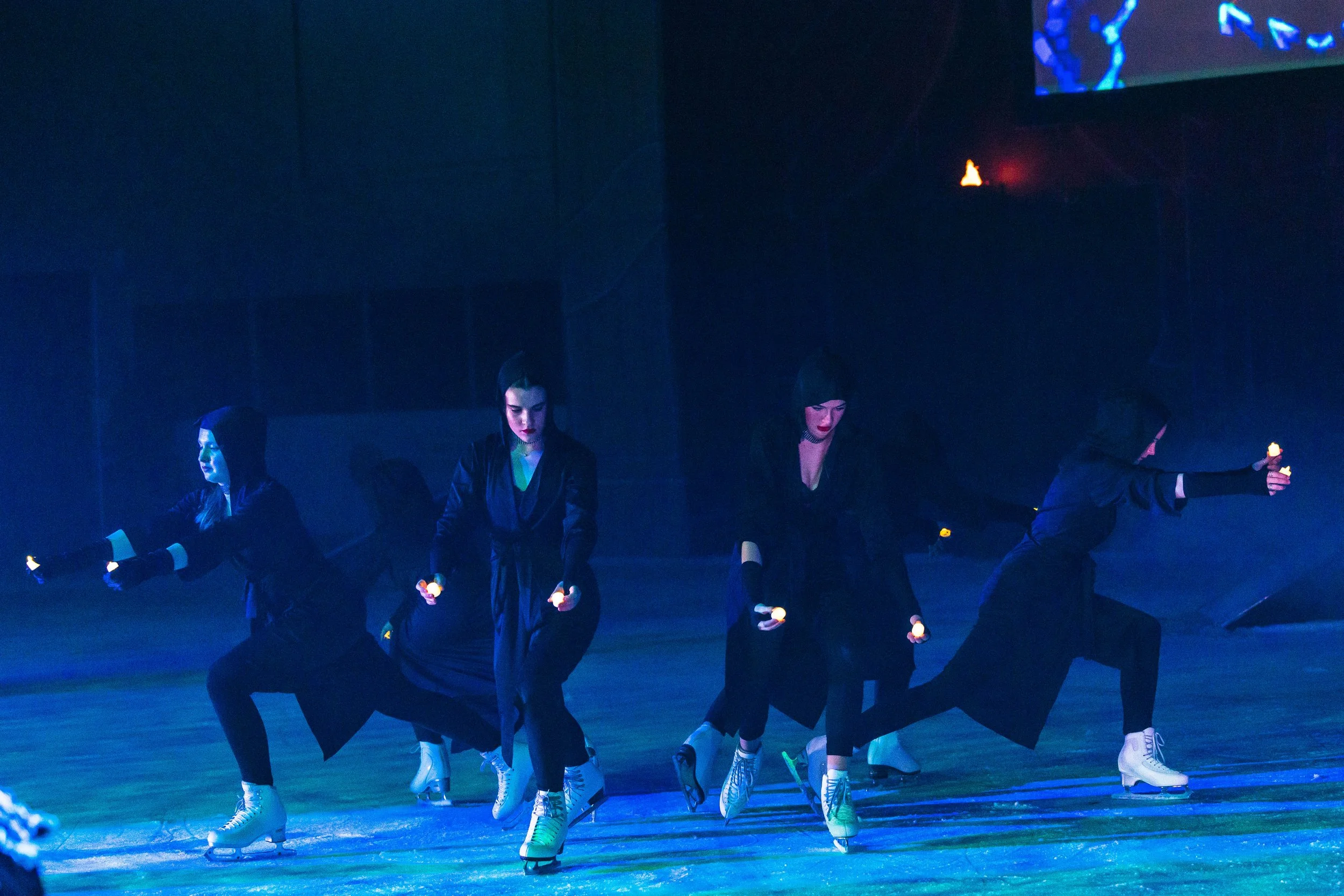 A group of five women ice skating indoors at night, holding small glowing objects while dressed in black clothing and ice skates.