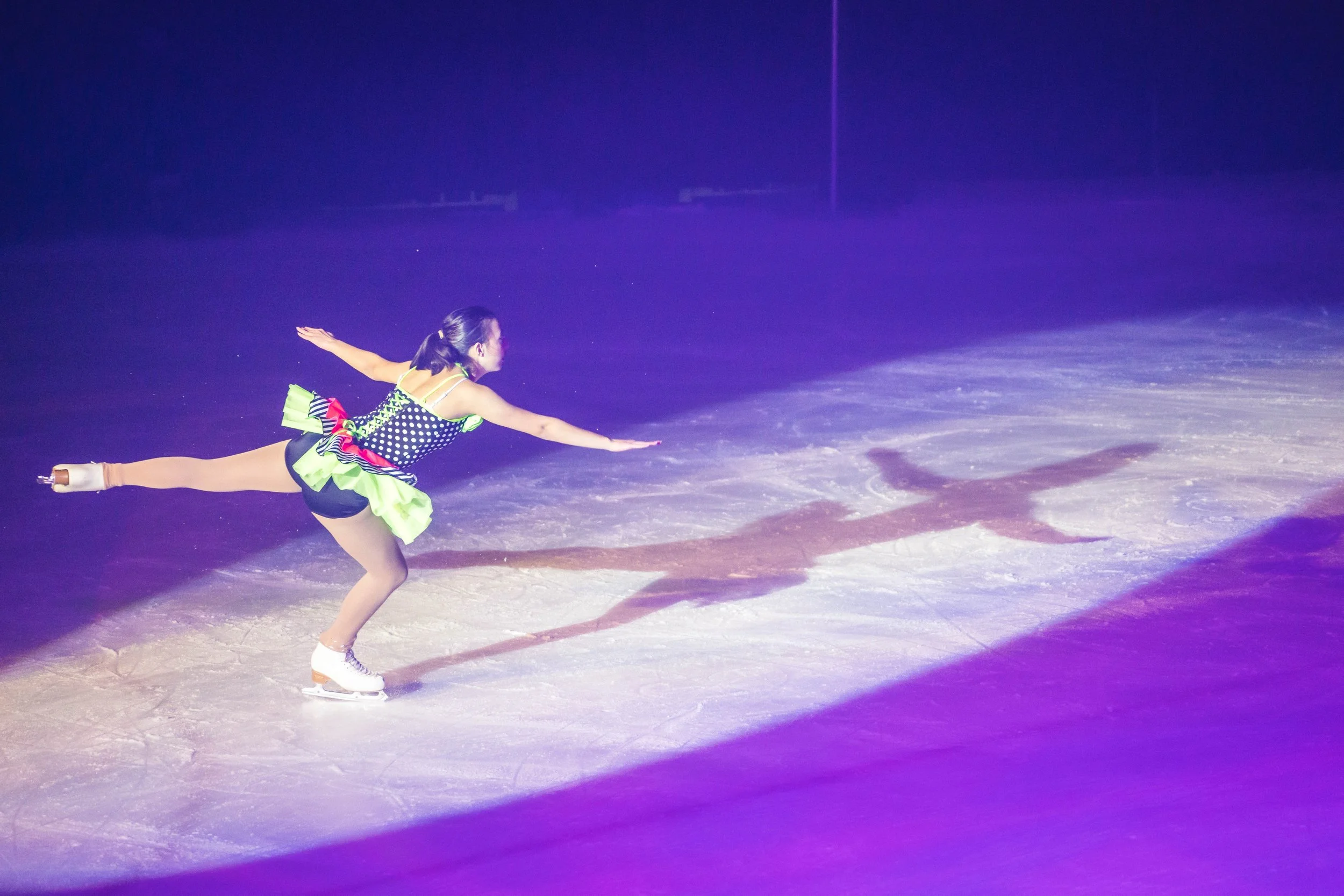 A female figure skater in a polka-dot costume is performing a spin on the ice rink under colorful lights.