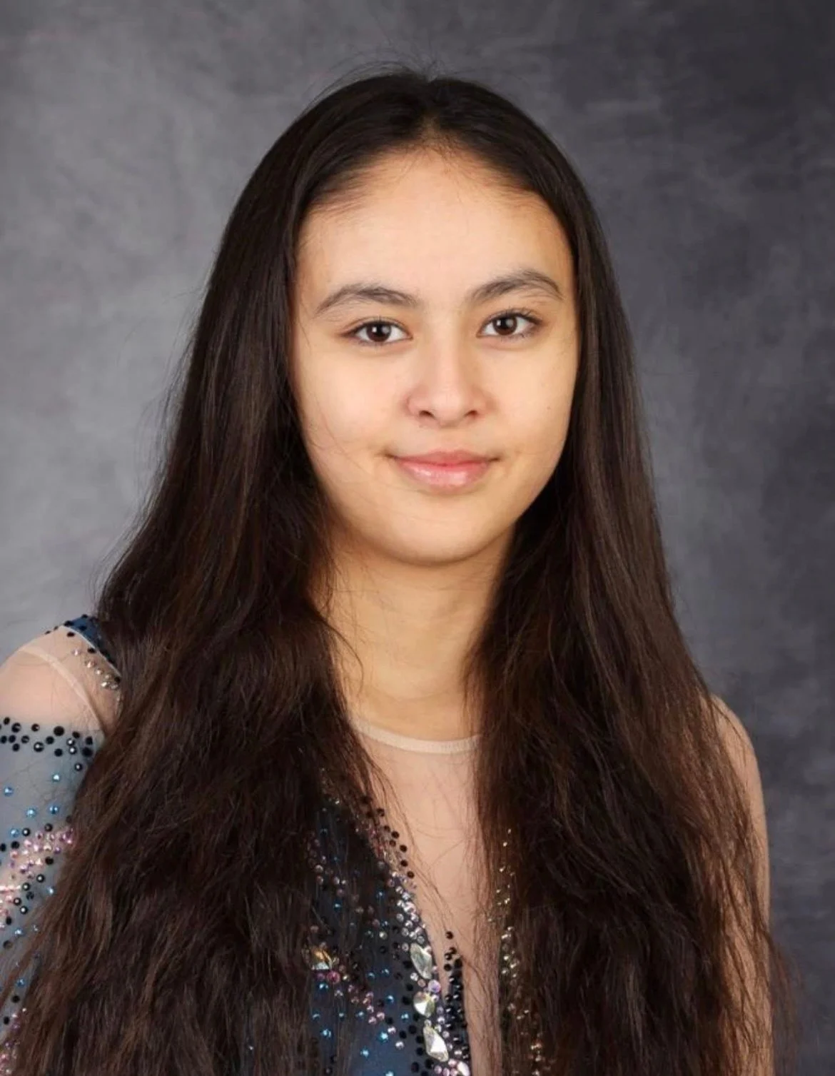 Young woman with long dark hair, wearing a sparkly dress, smiling softly against a gray background.
