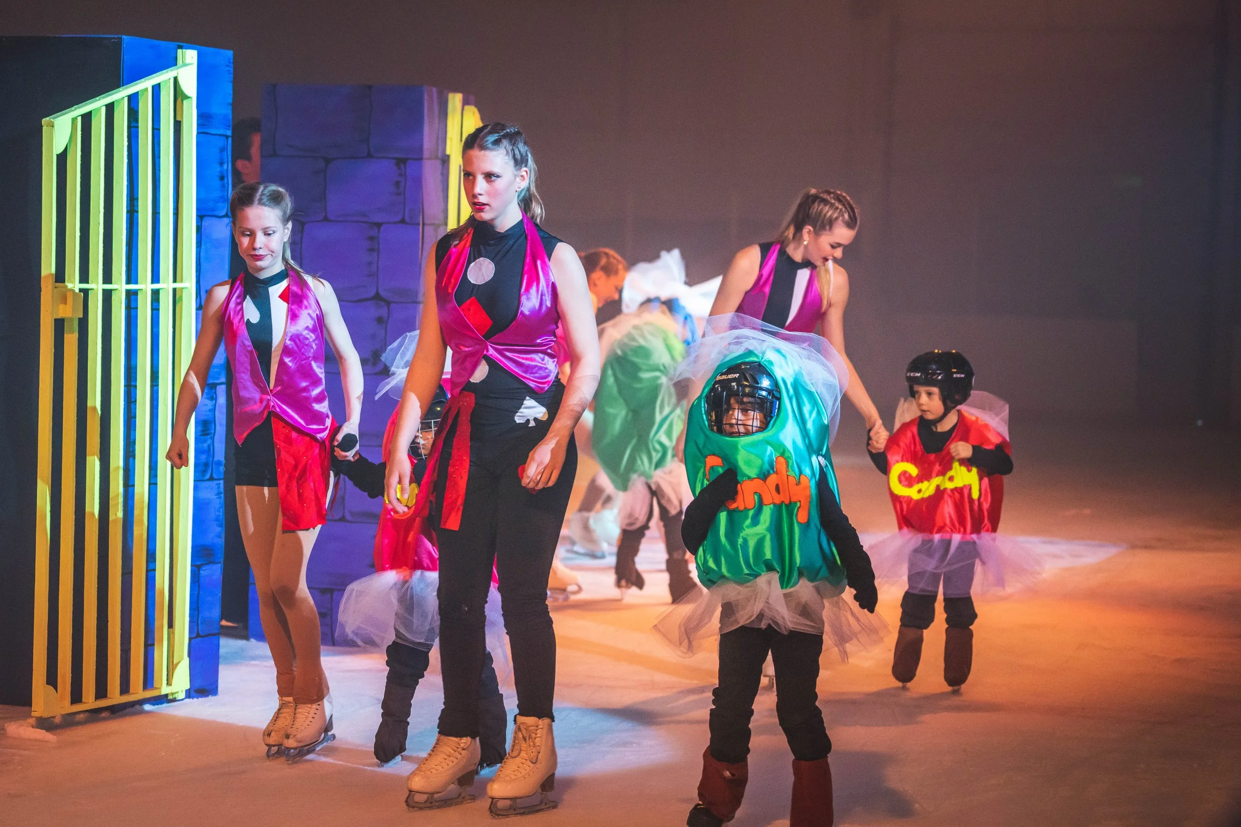 Children in colorful costumes performing a dance on stage, with some wearing hockey helmets and skating shoes.