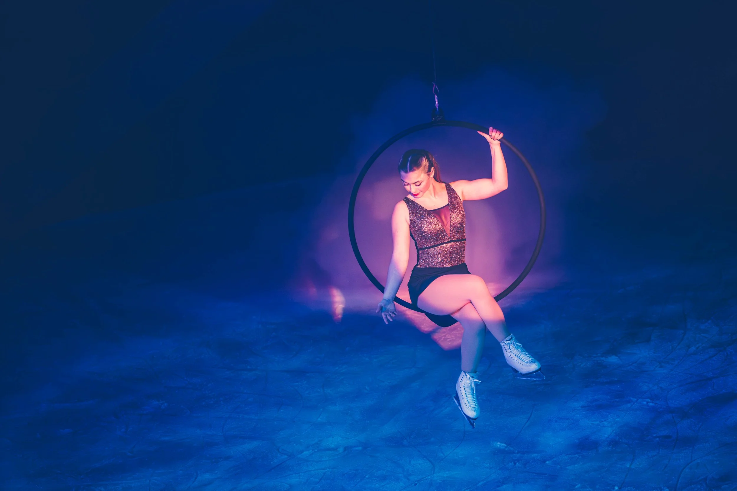 A female performer dressed in a sparkly black and maroon outfit sitting on the floor of a dark stage, holding a large hoop above her head with her right hand, surrounded by purple and blue lighting, ice skates on her feet.