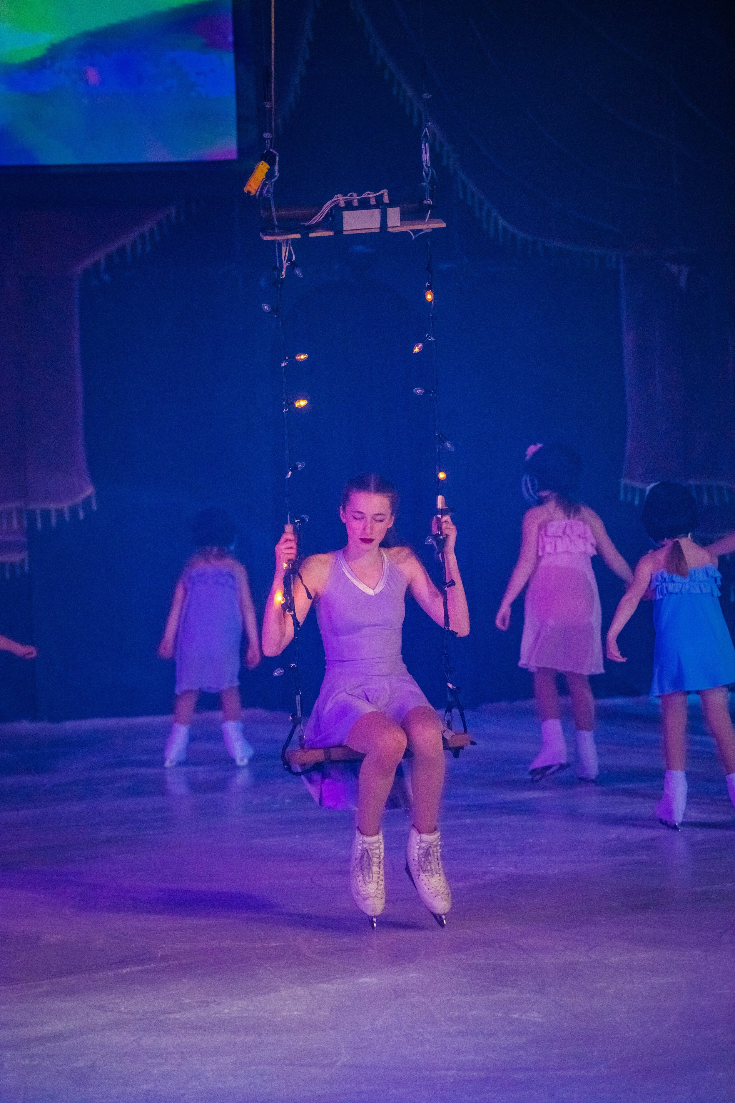A young woman in a ballet costume on ice skates is seated on a swing, with her hands holding the ropes of the swing. She is surrounded by other young ballerinas in pastel pink and blue costumes, some dancing or standing nearby on an ice rink, with a 