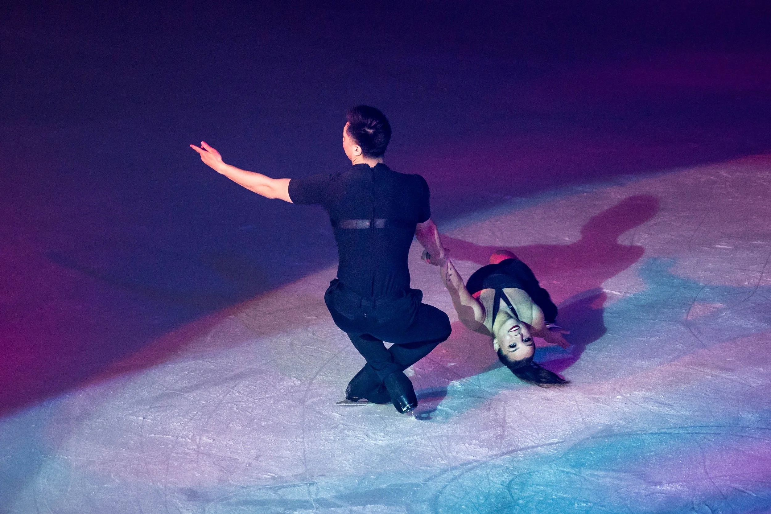 A figure skater in a pink costume performing a move on the ice rink with colorful stage lighting creating a vibrant atmosphere.