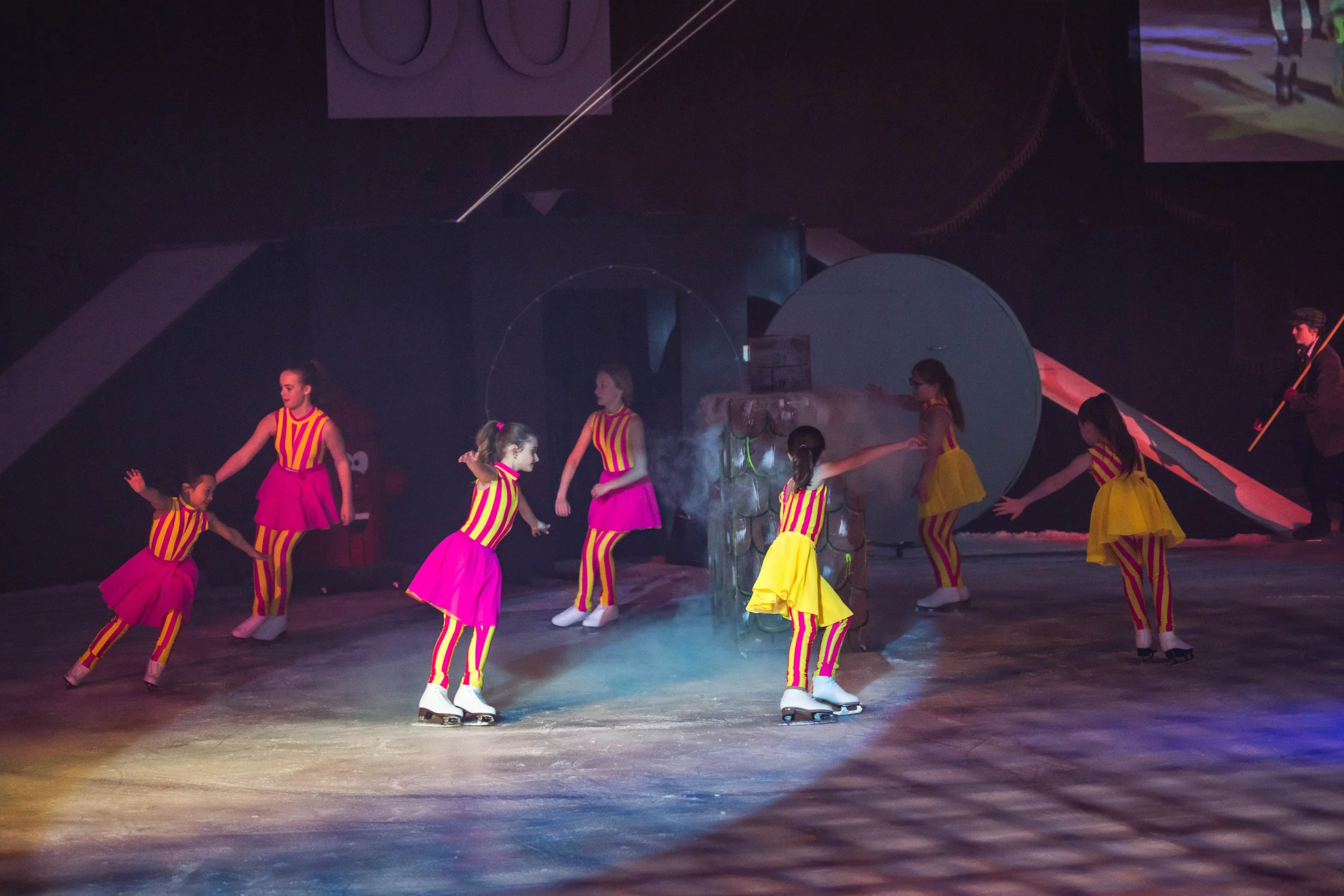 A group of young girls ice skating on an ice rink, wearing colorful striped costumes. Some are holding hands or skating in different directions, and there is a man in dark clothing holding a broom or stick nearby.