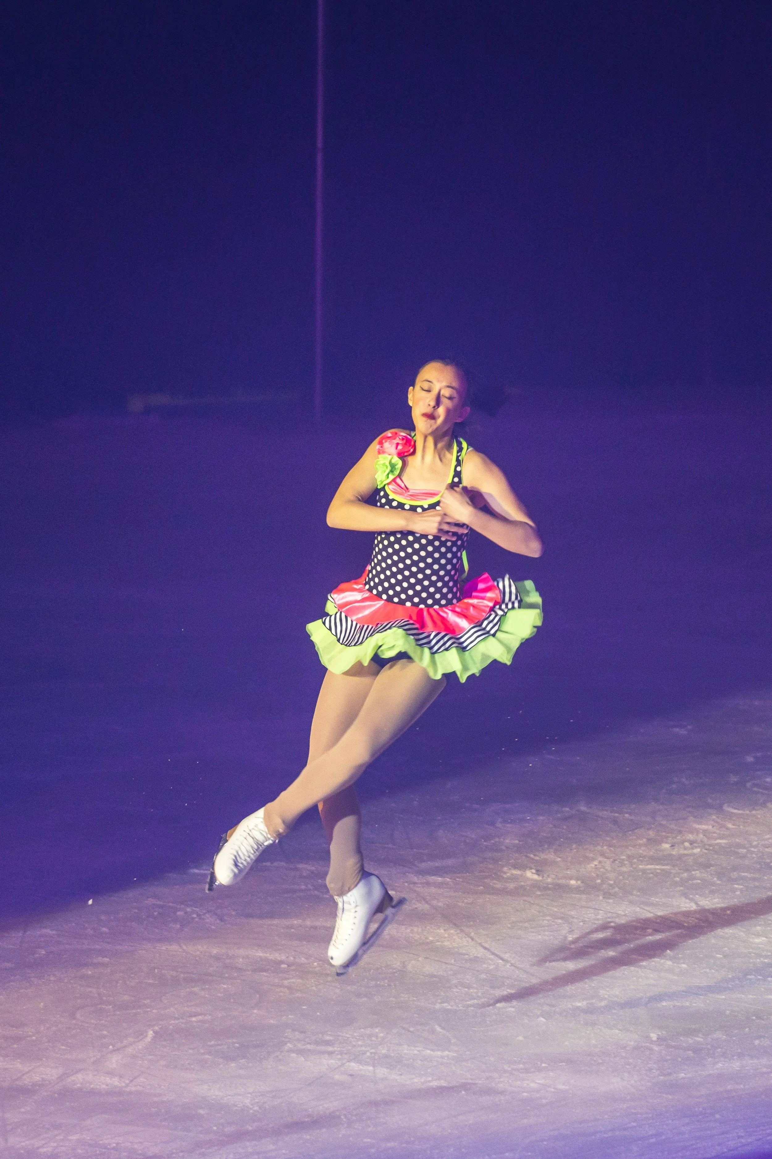 A female figure skater performing on ice, dressed in a colorful polka dot costume with a ruffled skirt, wearing white skates, against a dark background with purple lighting.