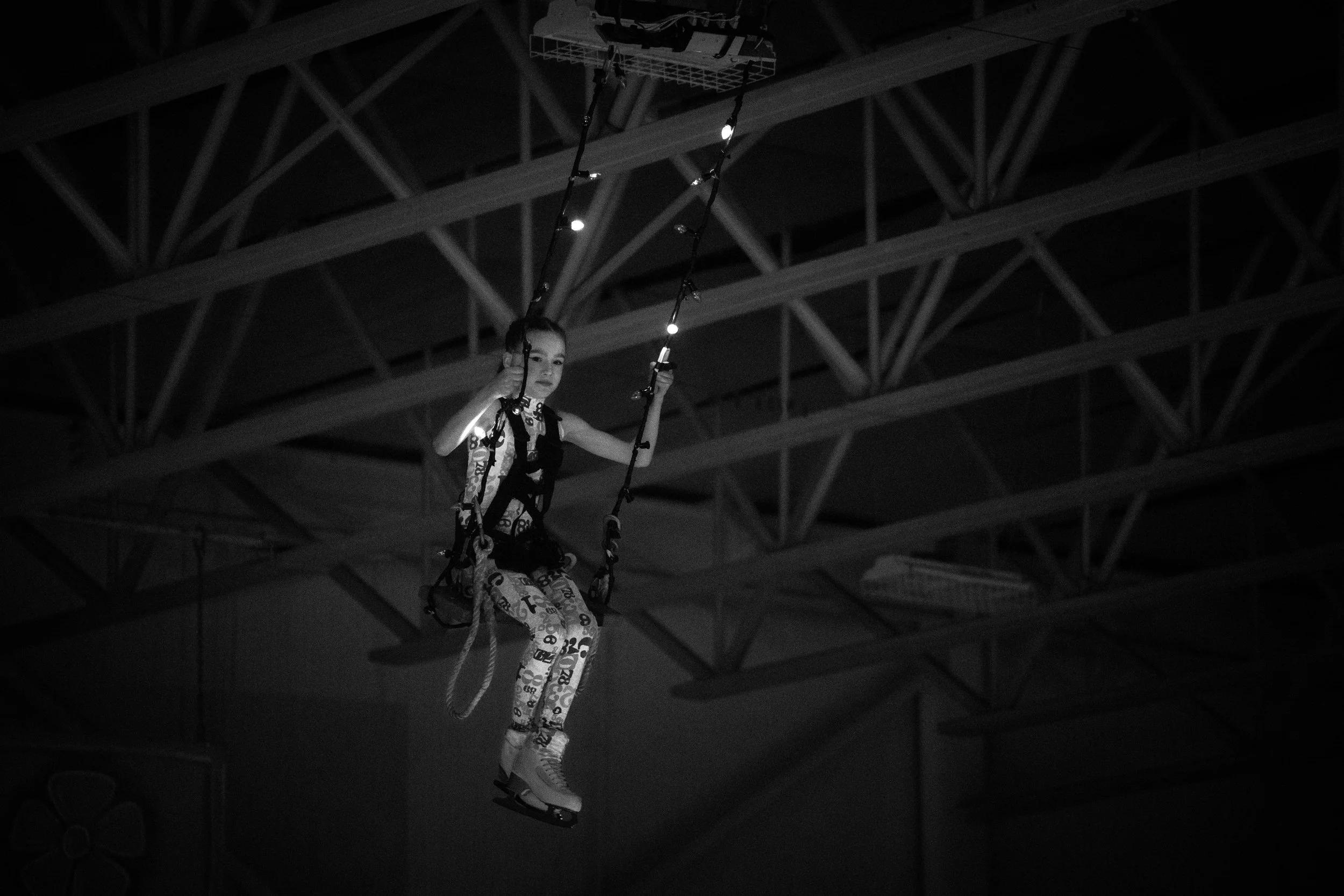 A young girl wearing patterned pants and high-top shoes is suspended in a harness at an indoor trampoline park, with a high ceiling and structural beams visible above her.