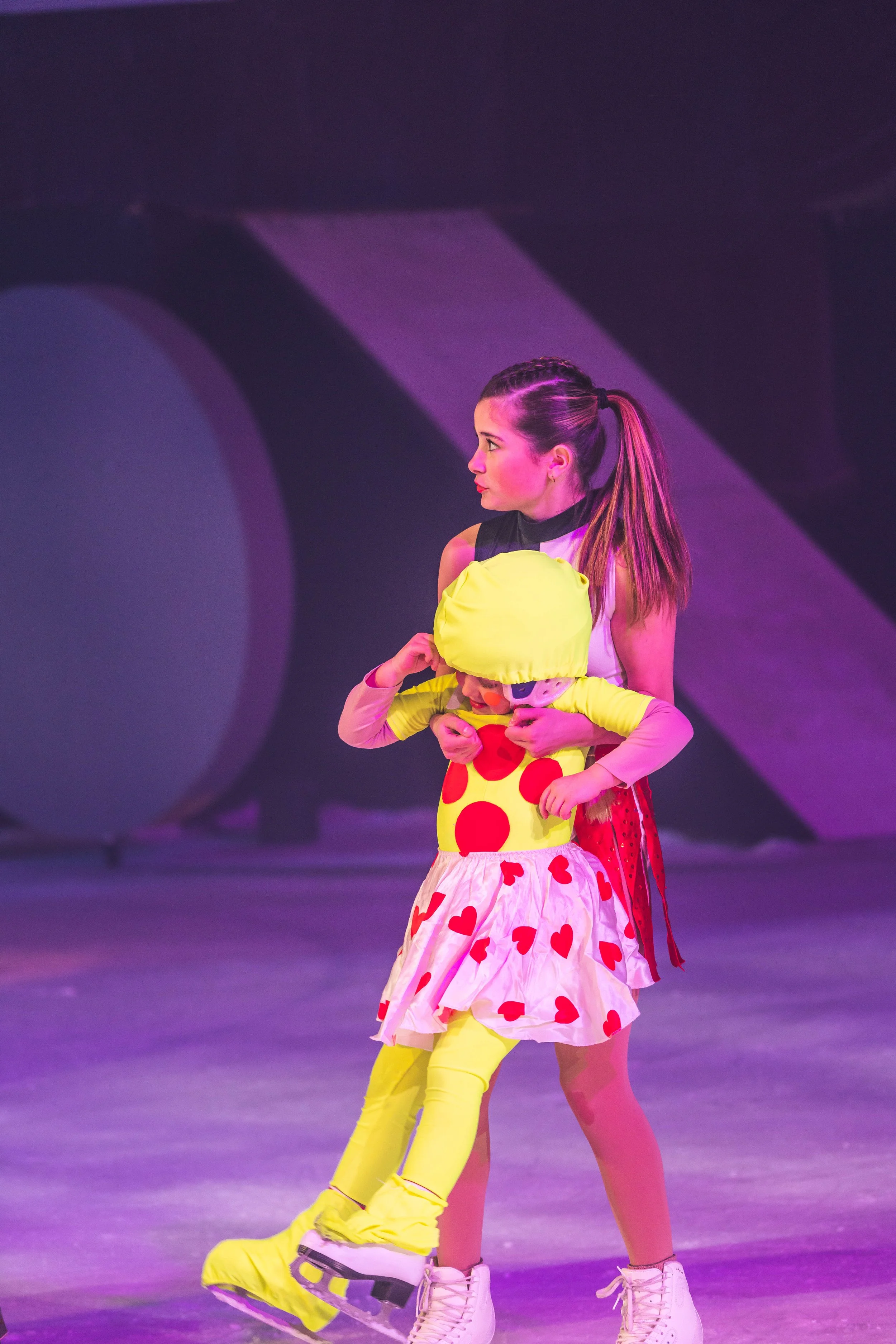 Two women ice skating on an ice rink, one holding a young girl. All are dressed in colorful, playful costumes with bright yellow and red, featuring hearts and polka dots. The background is dark with purple lighting.