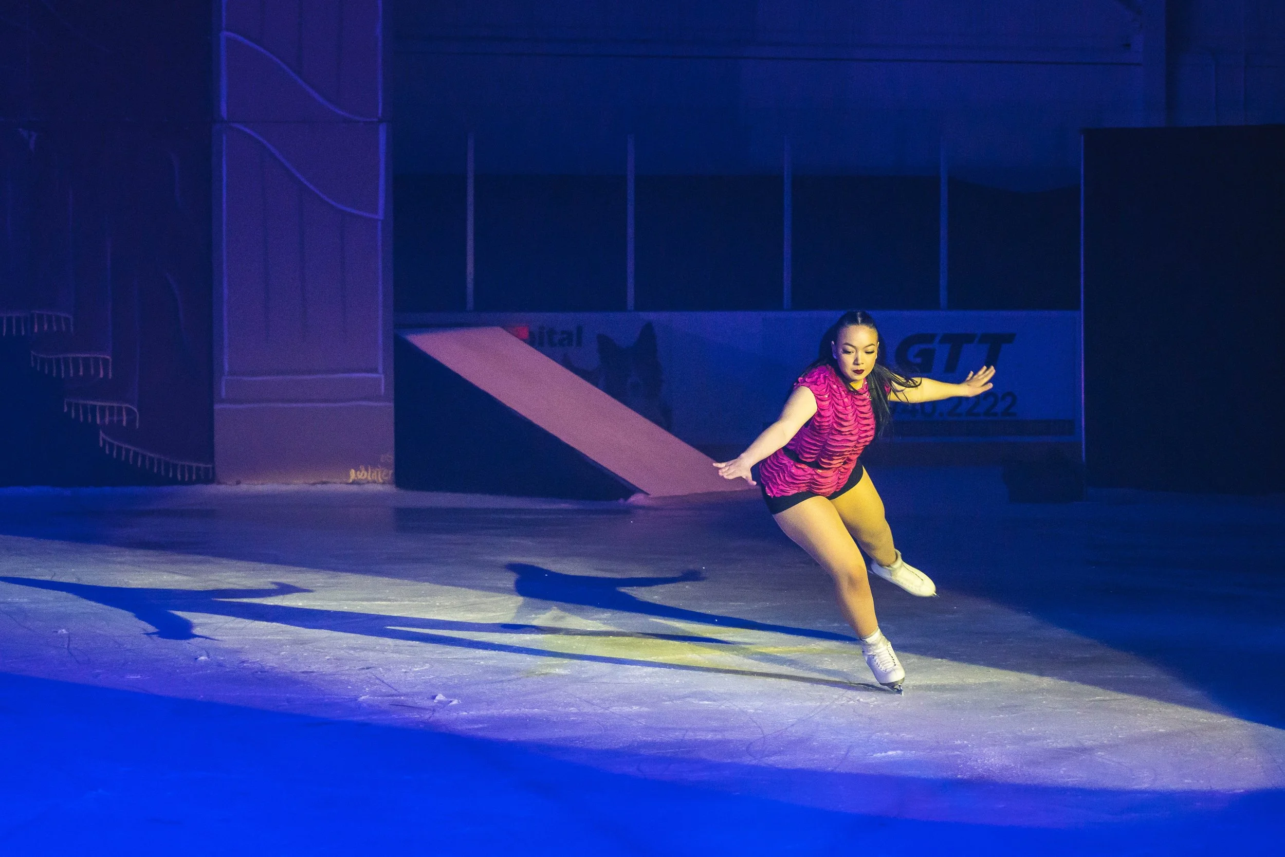 Figure skater in a pink and black costume performing on ice under blue stage lighting.