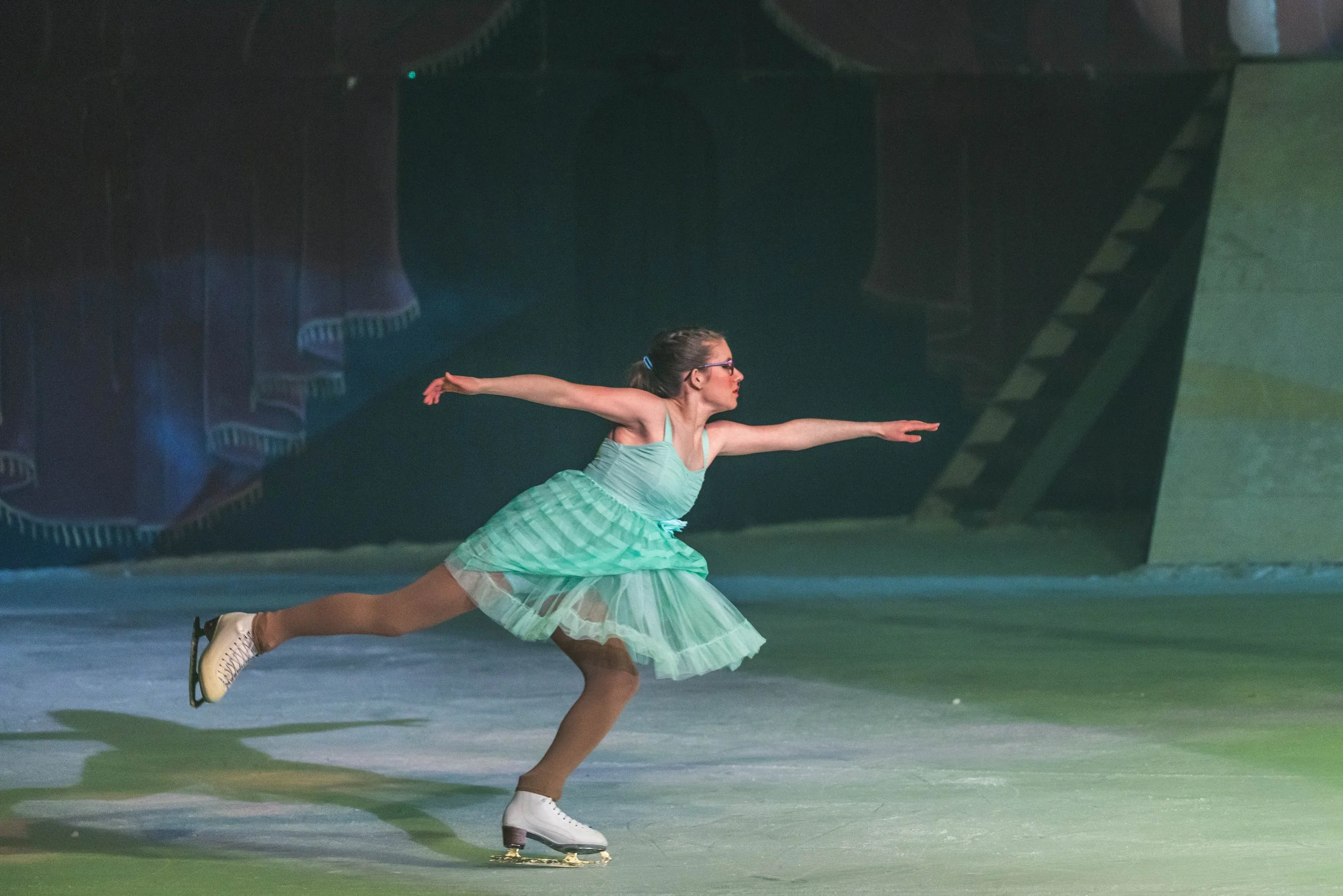 A female figure skater in a green dress performs a move on the ice, with her arms extended and one leg bent behind her. She wears glasses and ice skates, and the background features a stage with dark curtains and decorative elements.
