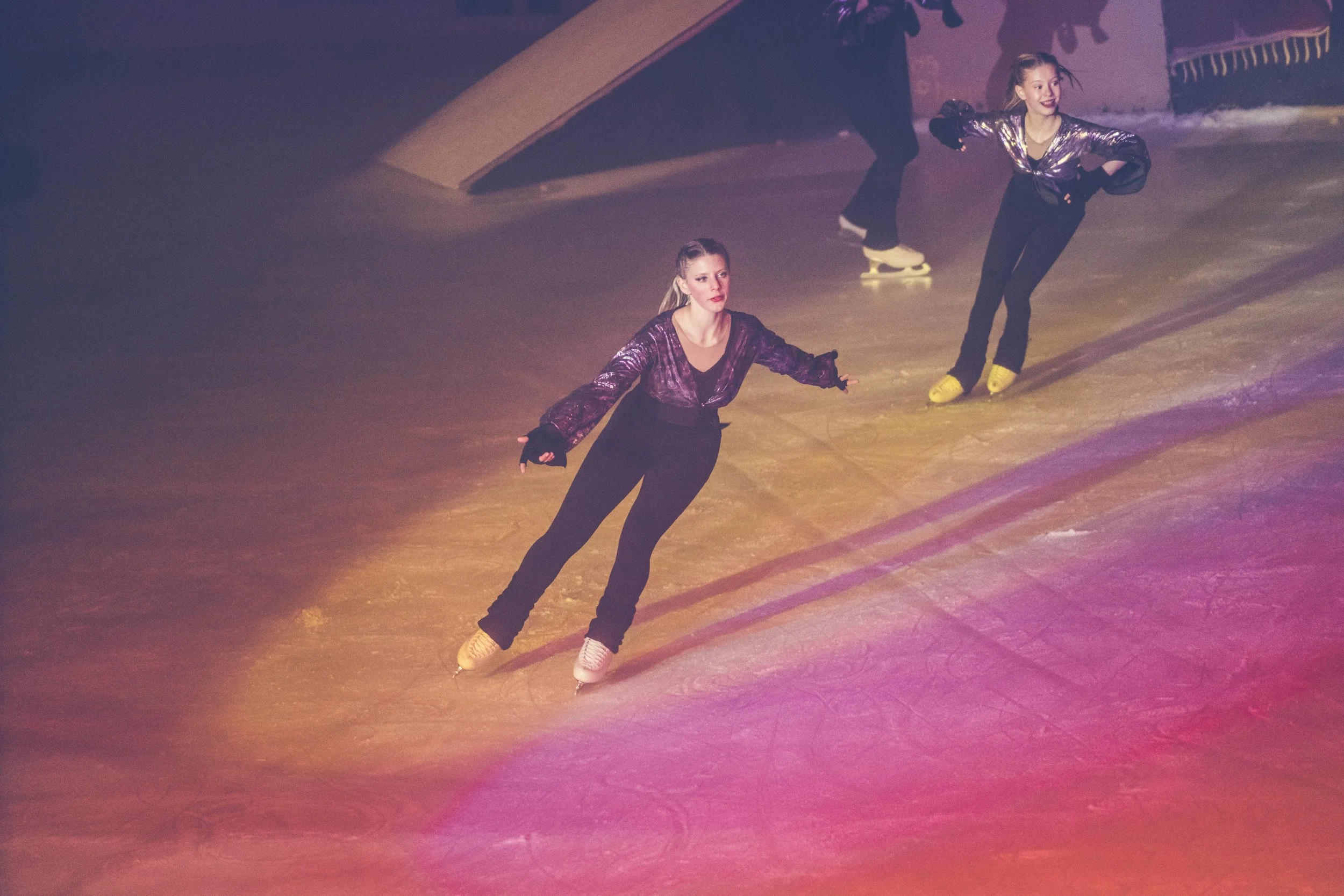 Two women ice skating on an indoor rink, wearing shiny jackets and black pants.