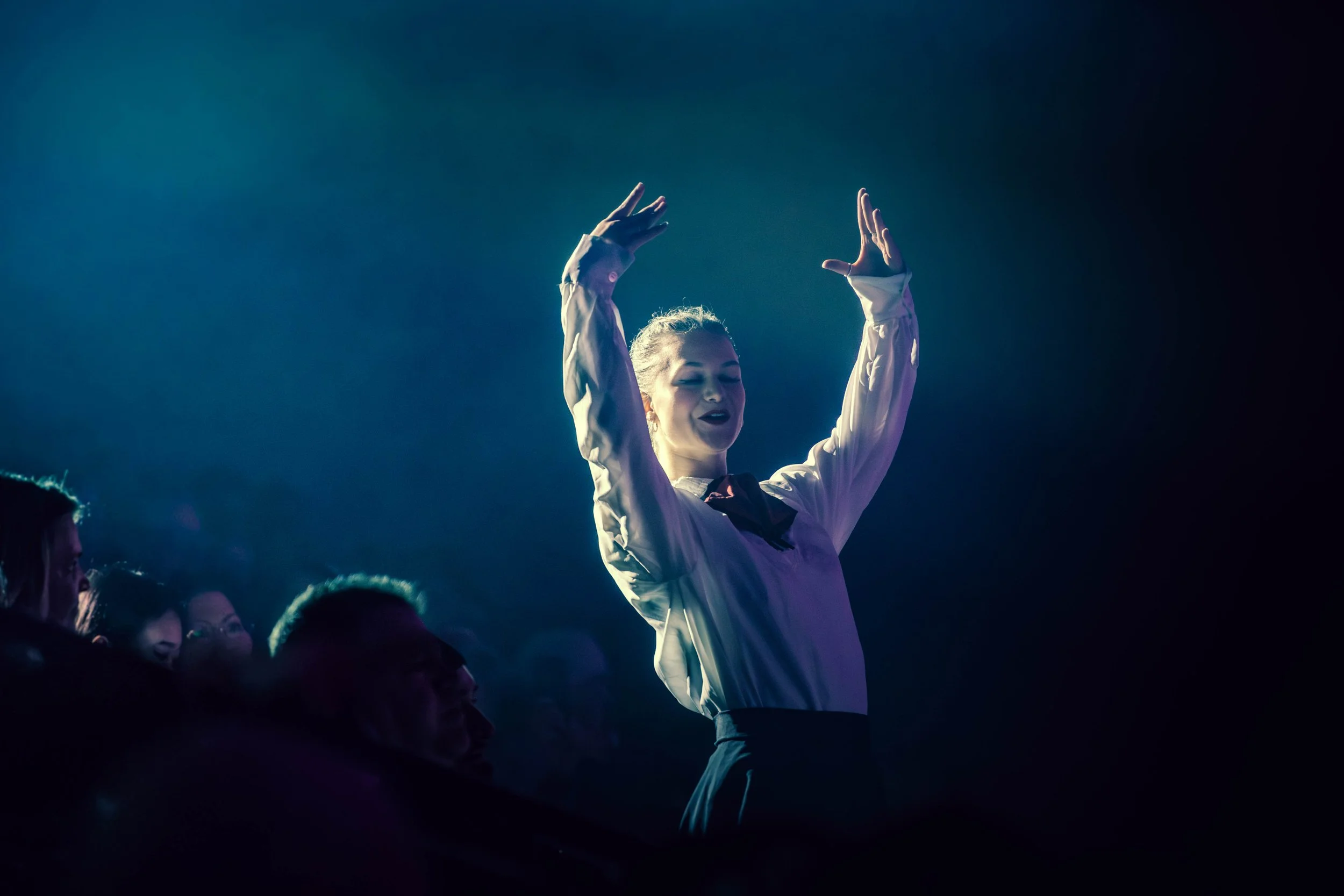 A woman with blonde hair, wearing a white blouse and a black tie, raises her hands in celebration amidst a dark background with blue stage lighting. She is surrounded by an audience, some of whom are visible in the lower part of the image.