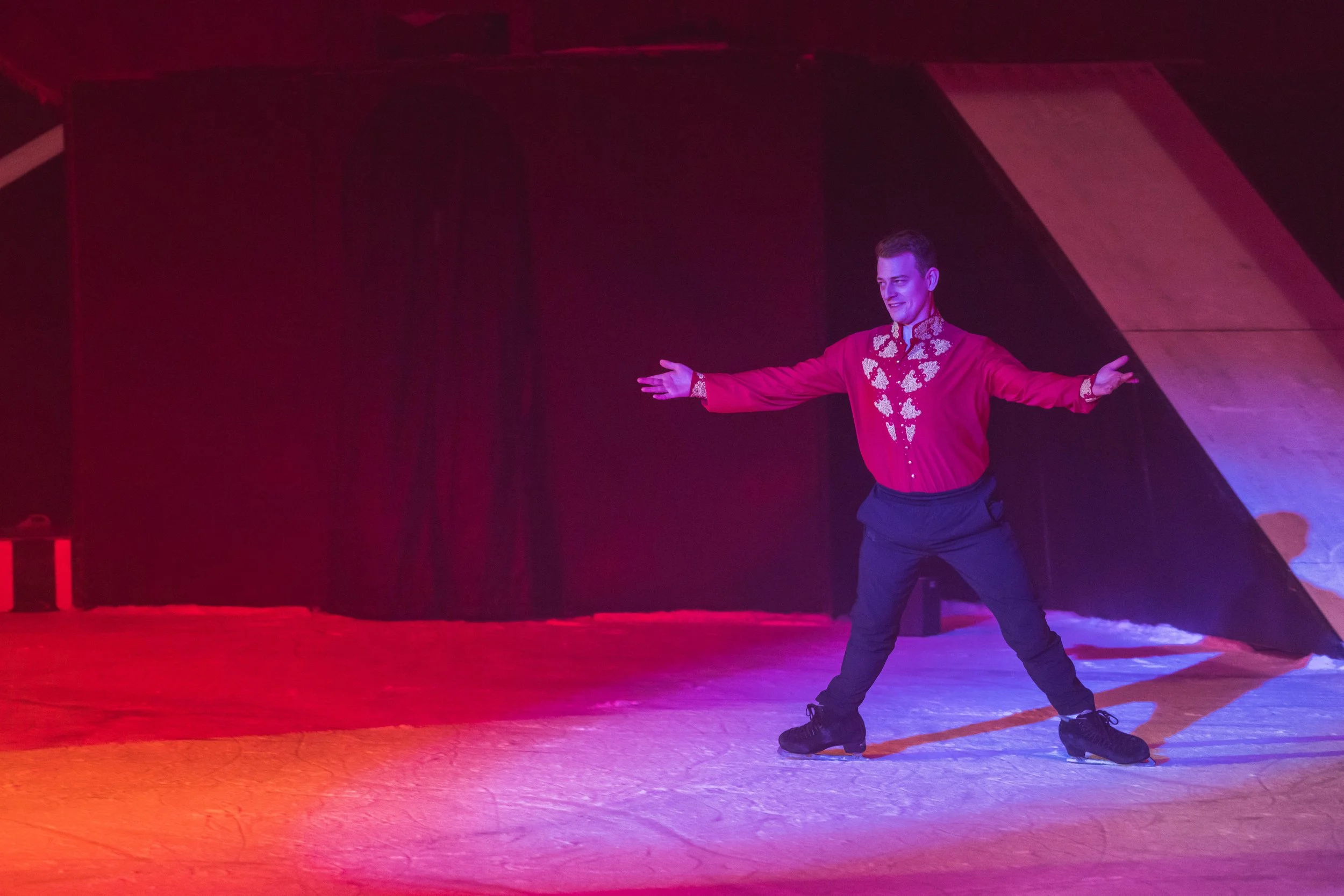 A man in a red traditional jacket with floral embroidery extending his arms while ice skating on an indoor rink.