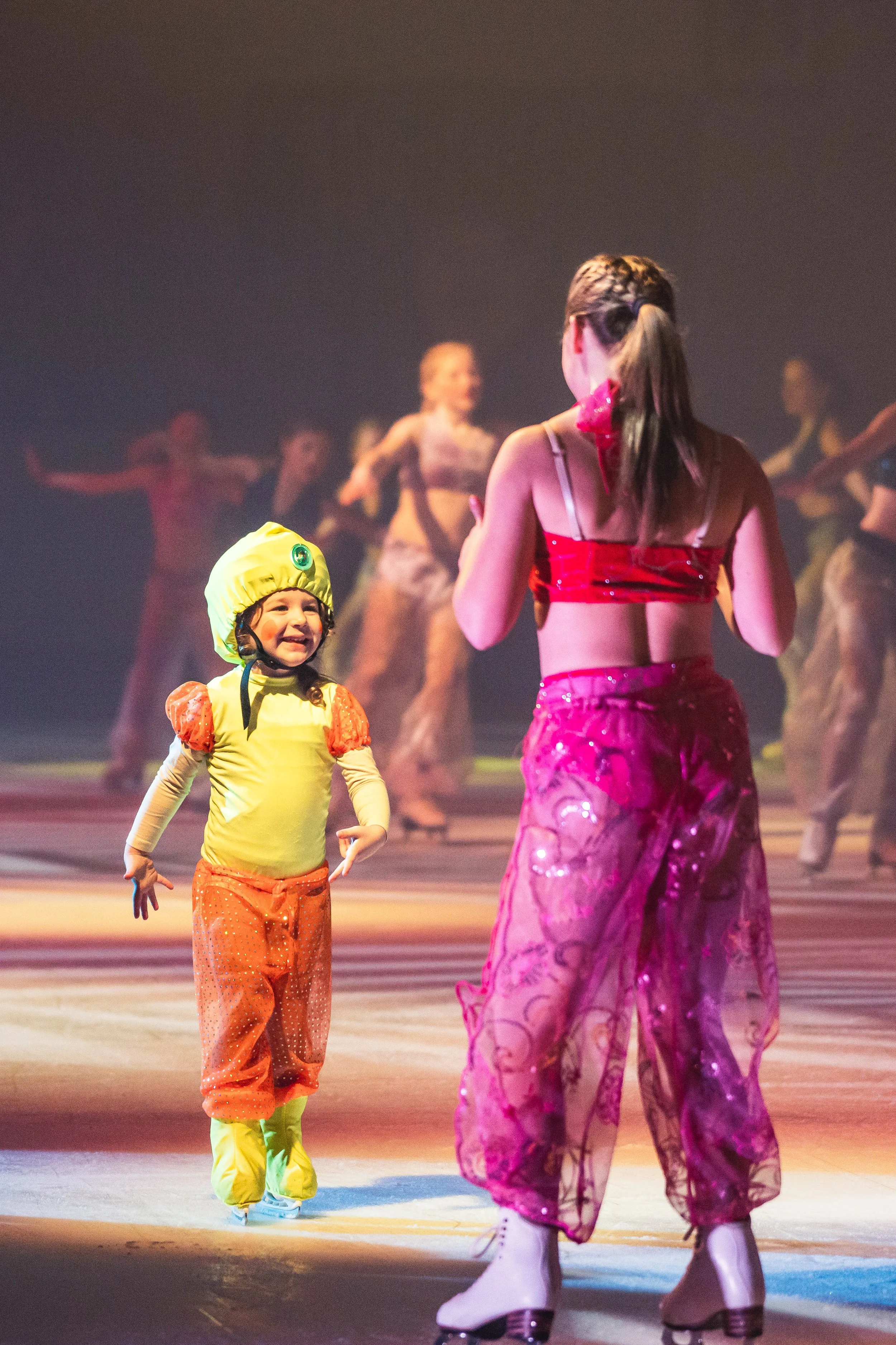 A young girl in an orange and yellow costume with a yellow hat and ice skate boots smiling at an adult wearing a bright pink and purple skirt with ice skates, with a background of other skaters on an ice rink.