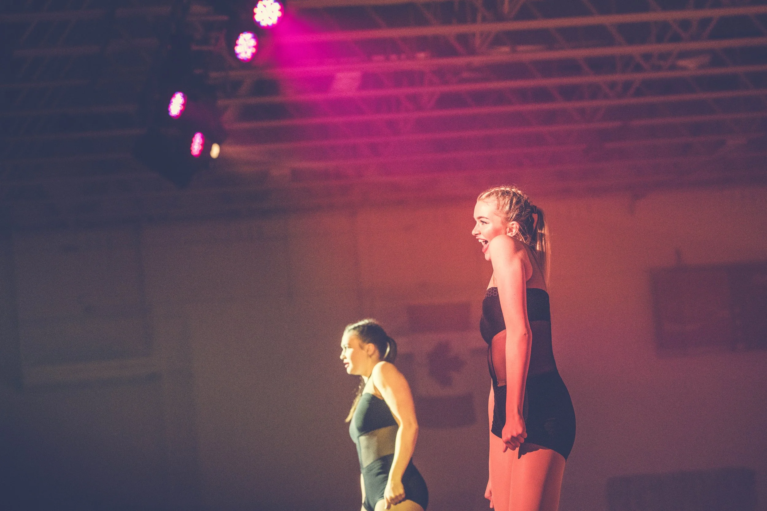 Three dancers are performing on a stage under colorful pink and purple lighting. They are in a line, smiling and looking forward, wearing dance costumes.