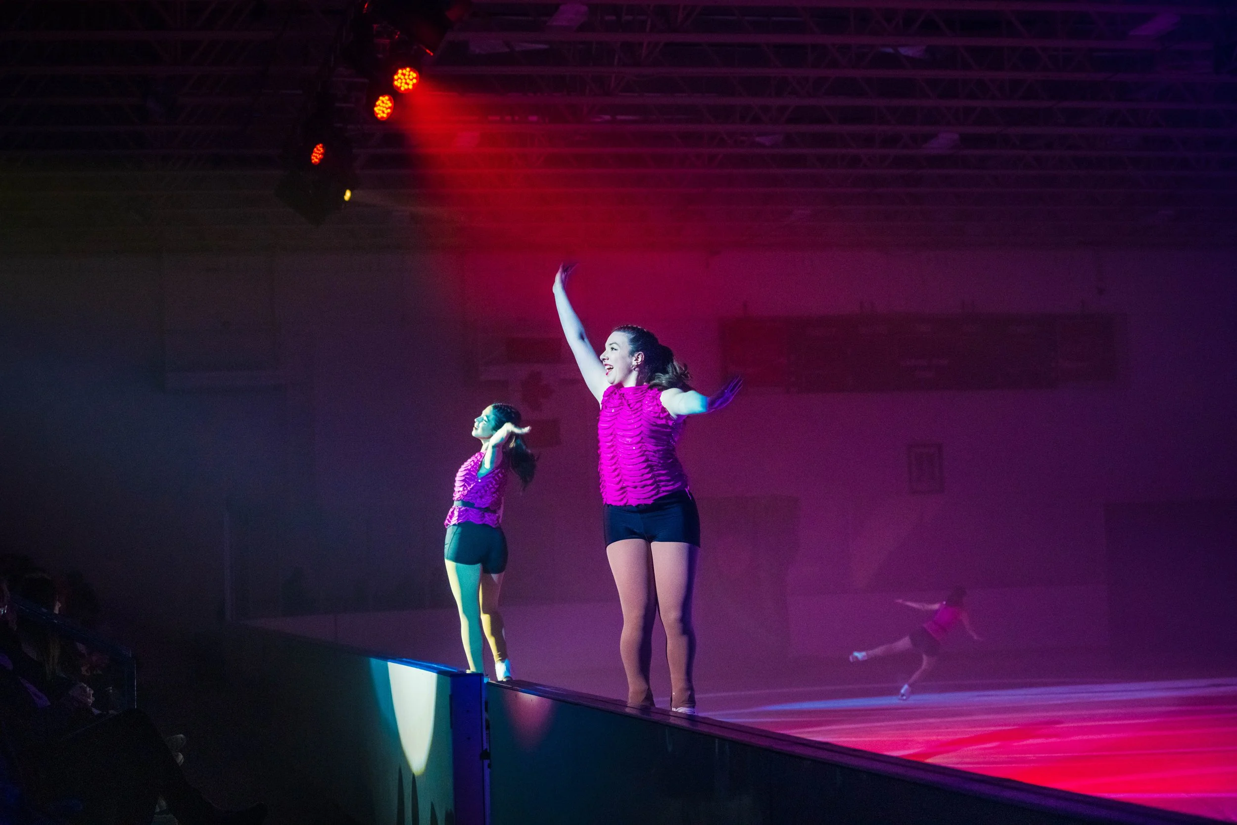 Two female performers in pink and purple costumes cheer on stage with arms raised, under purple and red stage lighting.