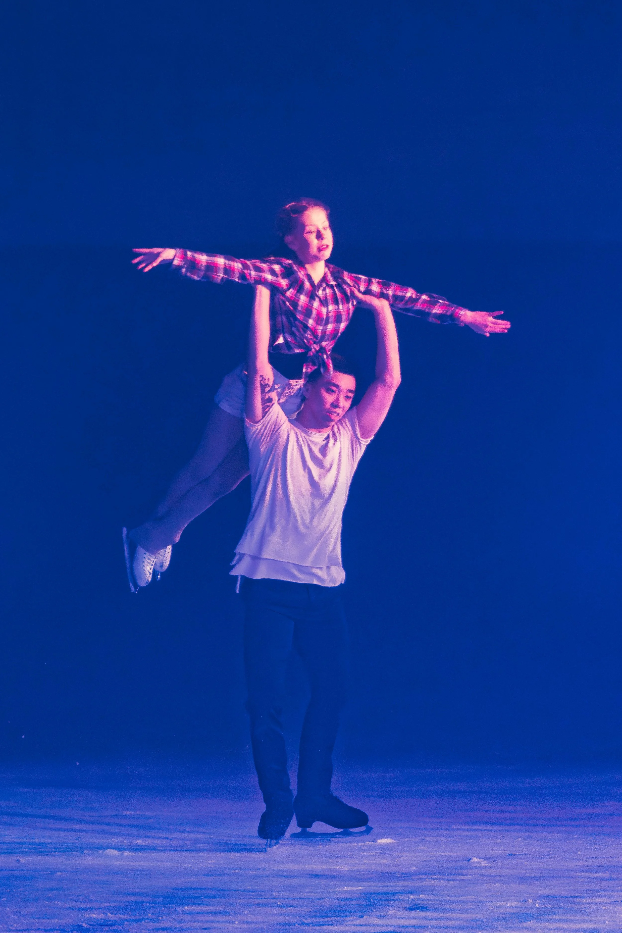 Two people are ice skating indoors, one person is standing on ice with arms raised, and the other person is riding on the person's shoulders with arms outstretched, wearing a plaid shirt and a white t-shirt.