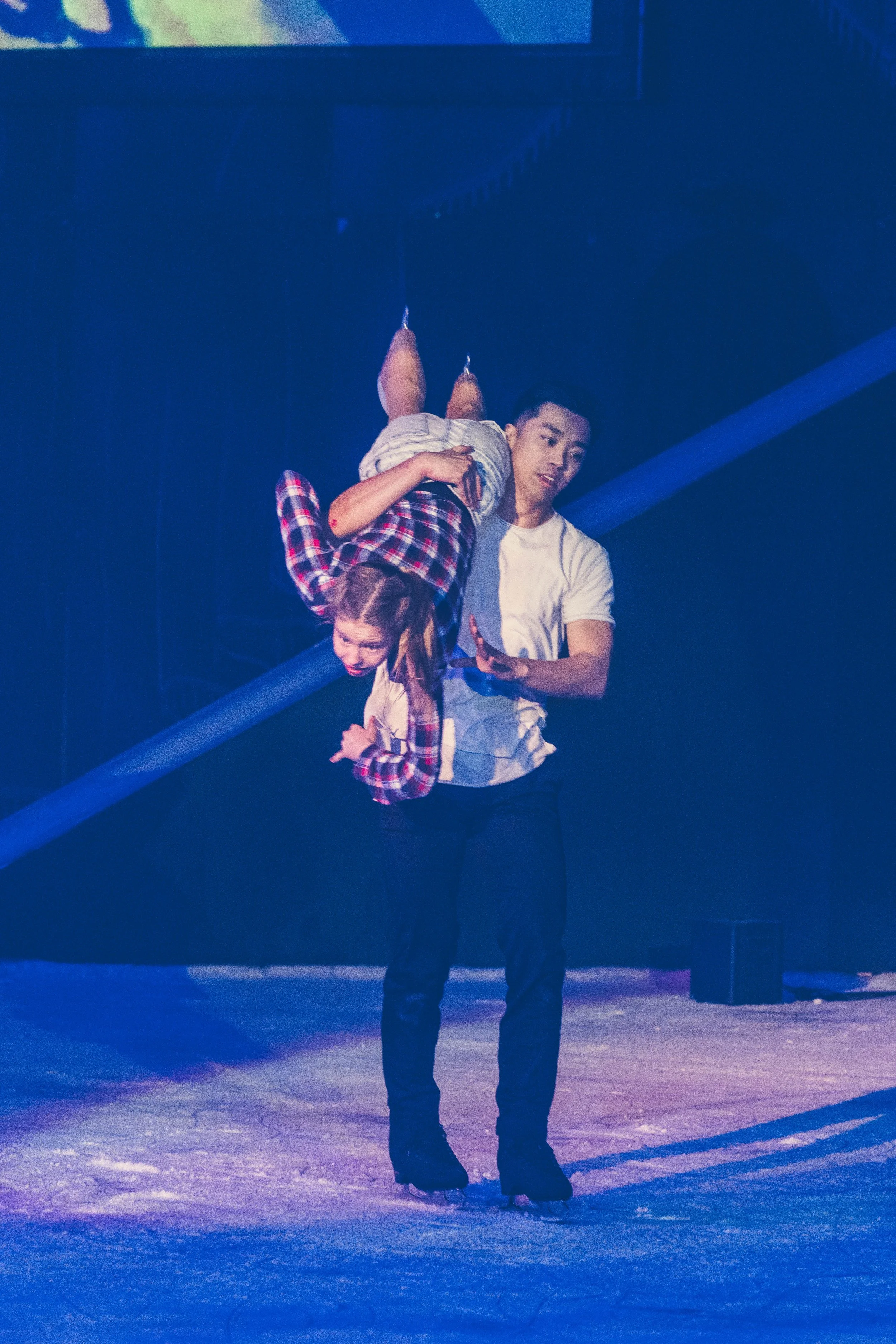Two people are ice skating, with one person holding the other upside down by the waist. The scene is illuminated with blue lighting and the ice surface has a shimmering appearance.