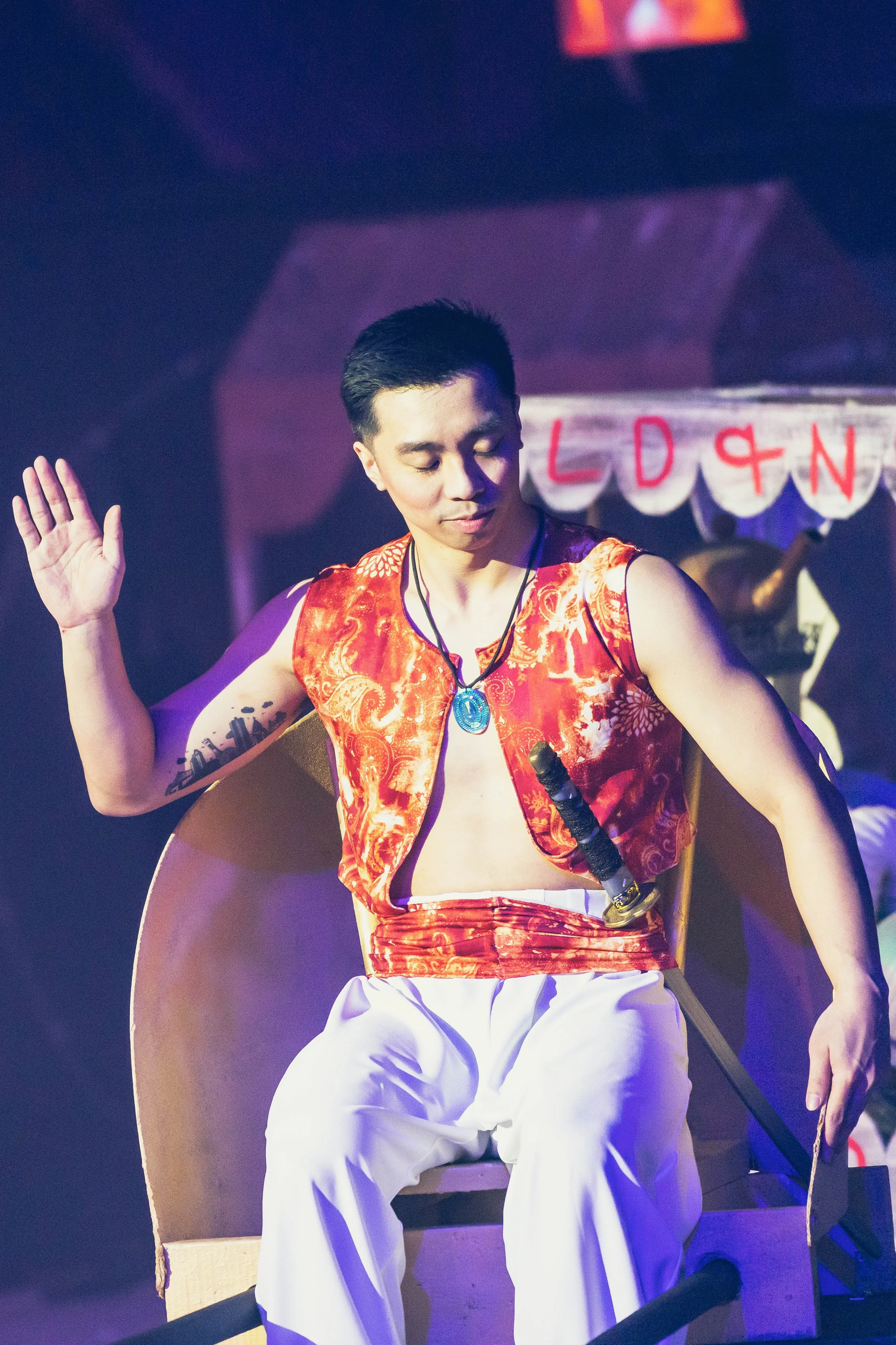 A person sitting on a stage with their eyes closed, wearing a red patterned vest, white pants, and a necklace with a blue pendant. Their right hand is raised, and they are holding a drumstick in their left hand. There is a person in the background ho