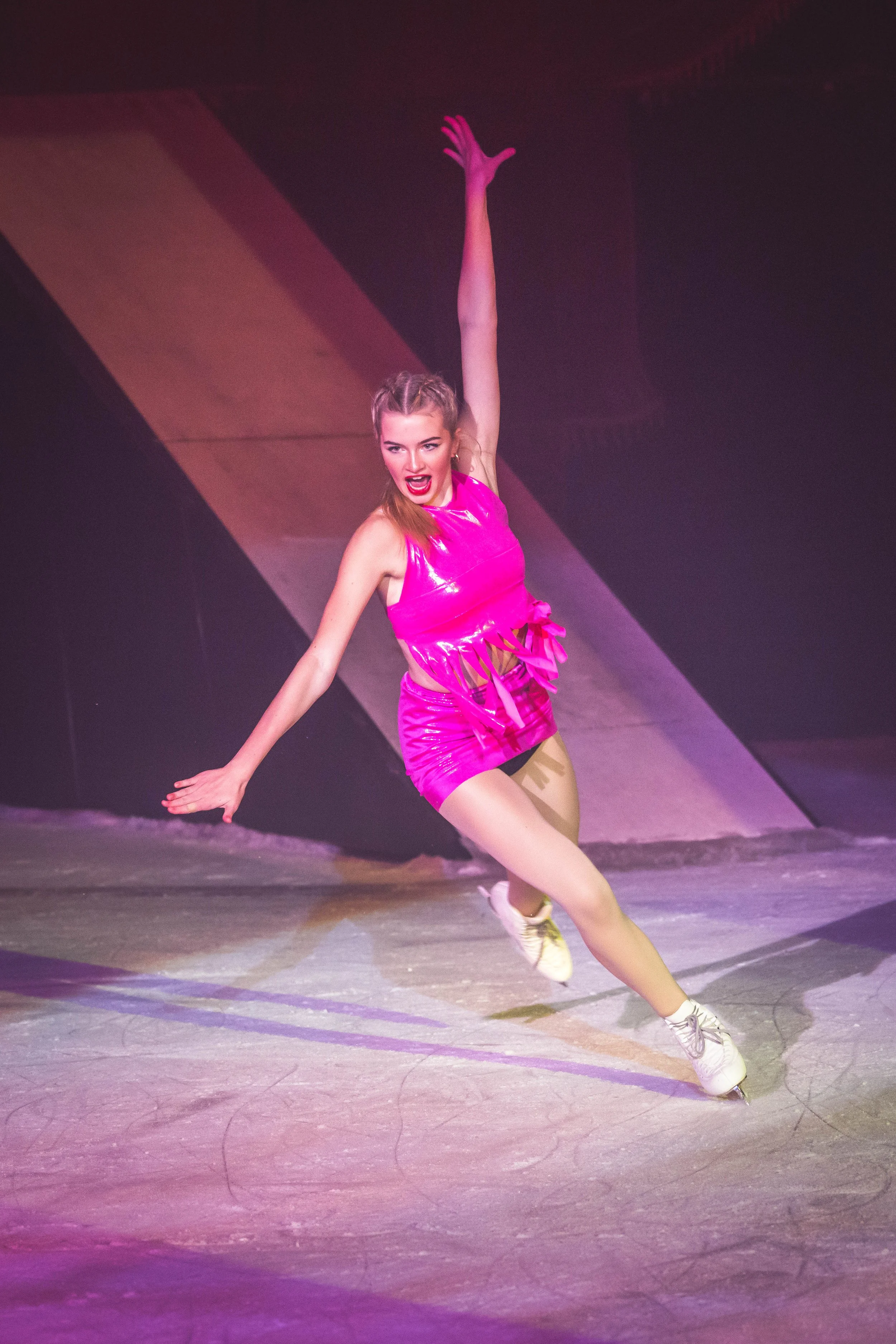 Young woman figure skating on ice rink wearing a bright pink costume, with her right arm raised and left arm extended to the side, performing a dance move.