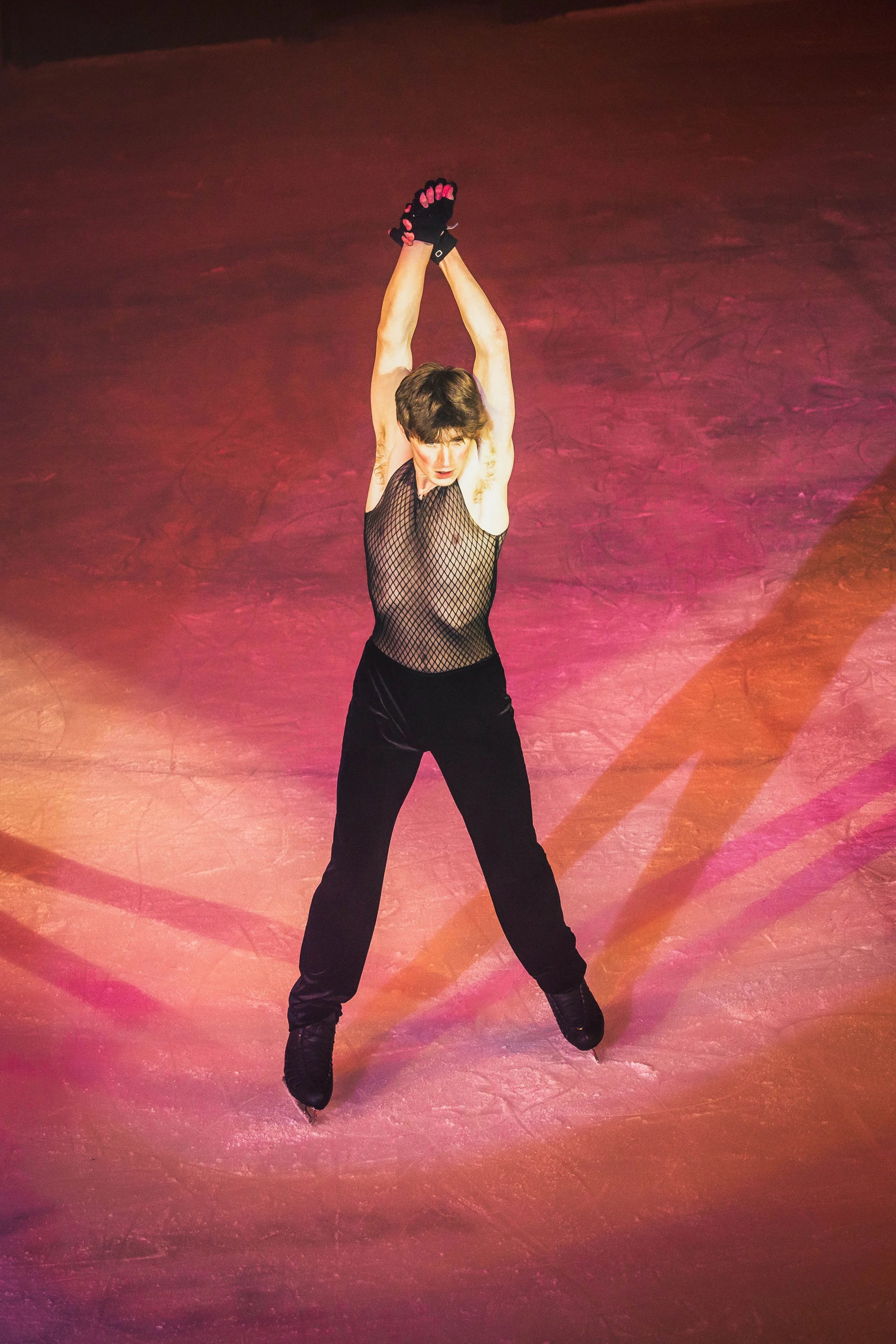A female figure skater wearing a black fishnet sleeveless top, black gloves, and black pants skating on an ice rink with pink and purple lighting.