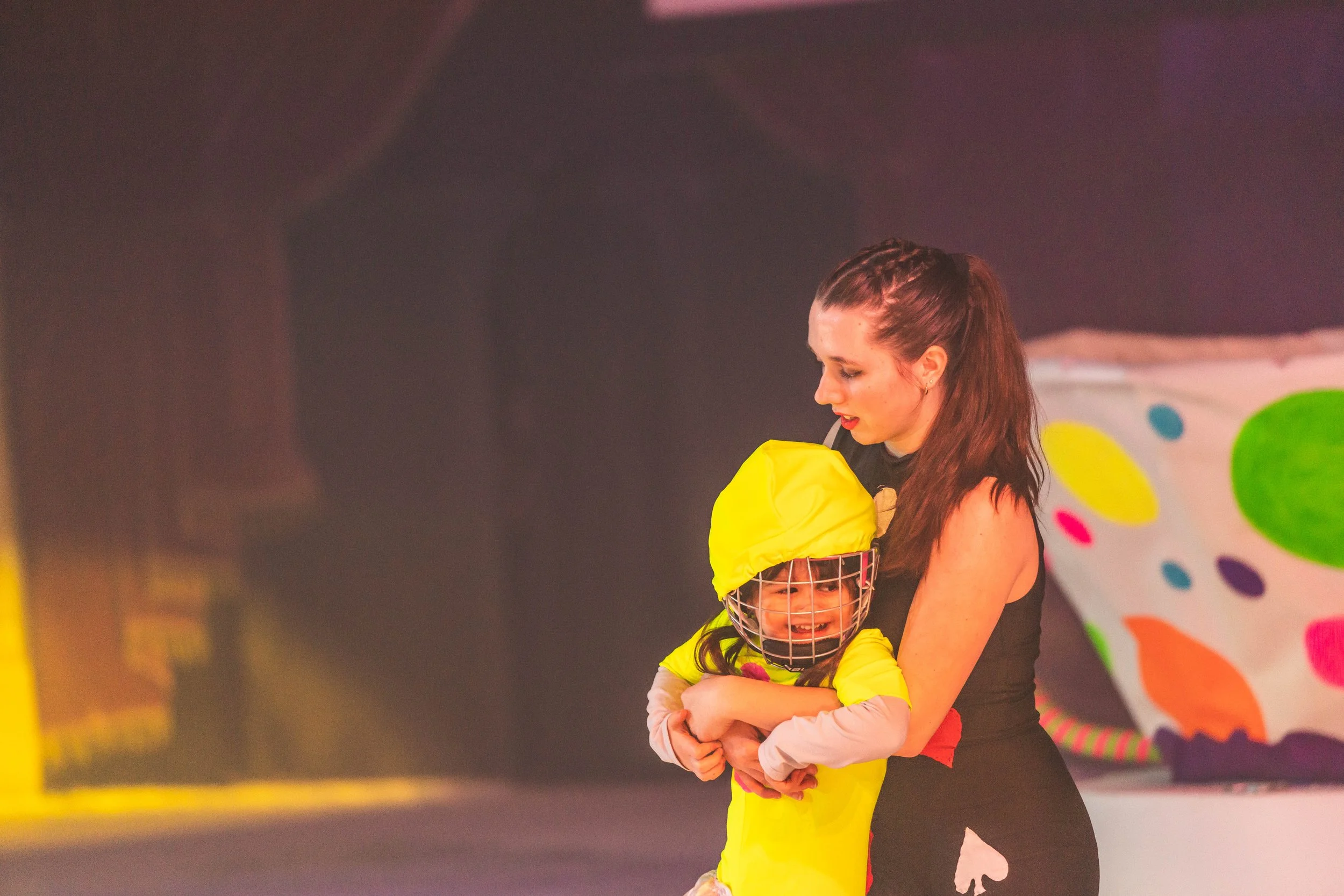 A young girl with curly hair, wearing a yellow helmet and a yellow outfit, is hugging a woman with long brown hair, who is wearing a black shirt. They are on a colorful stage or attraction.