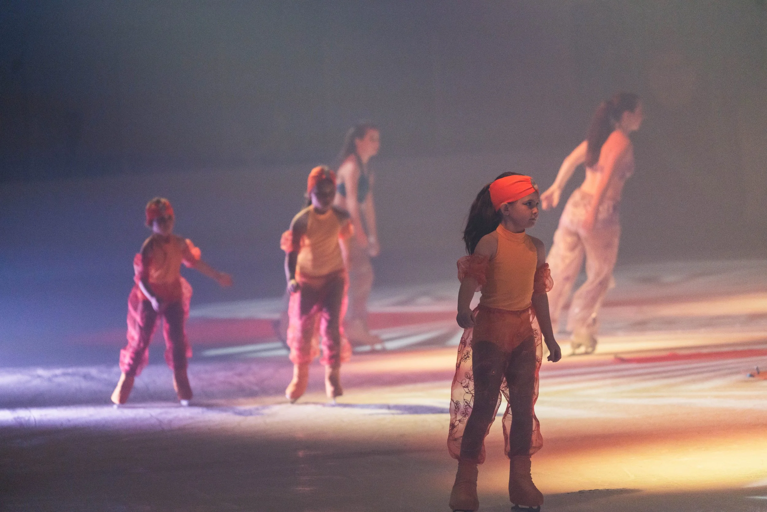 A group of children and an adult, possibly dancers, standing on a stage with atmospheric lighting. The children are dressed in colorful outfits with orange and red hues, some wearing headscarves, and appear to be preparing for a performance.