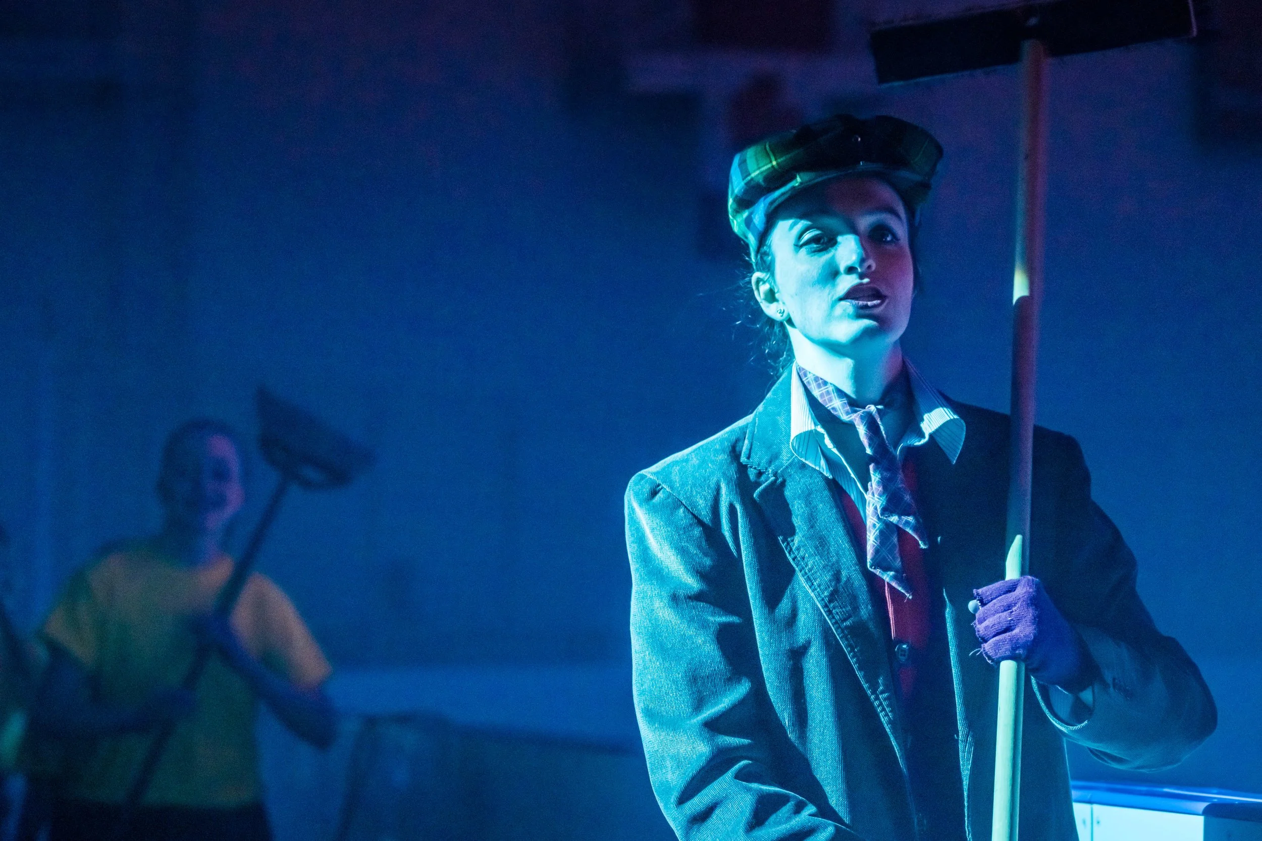 A woman with a plaid hat and a blue blazer holding a white cane, with another person blurred in the background, in a dark setting with blue lighting.