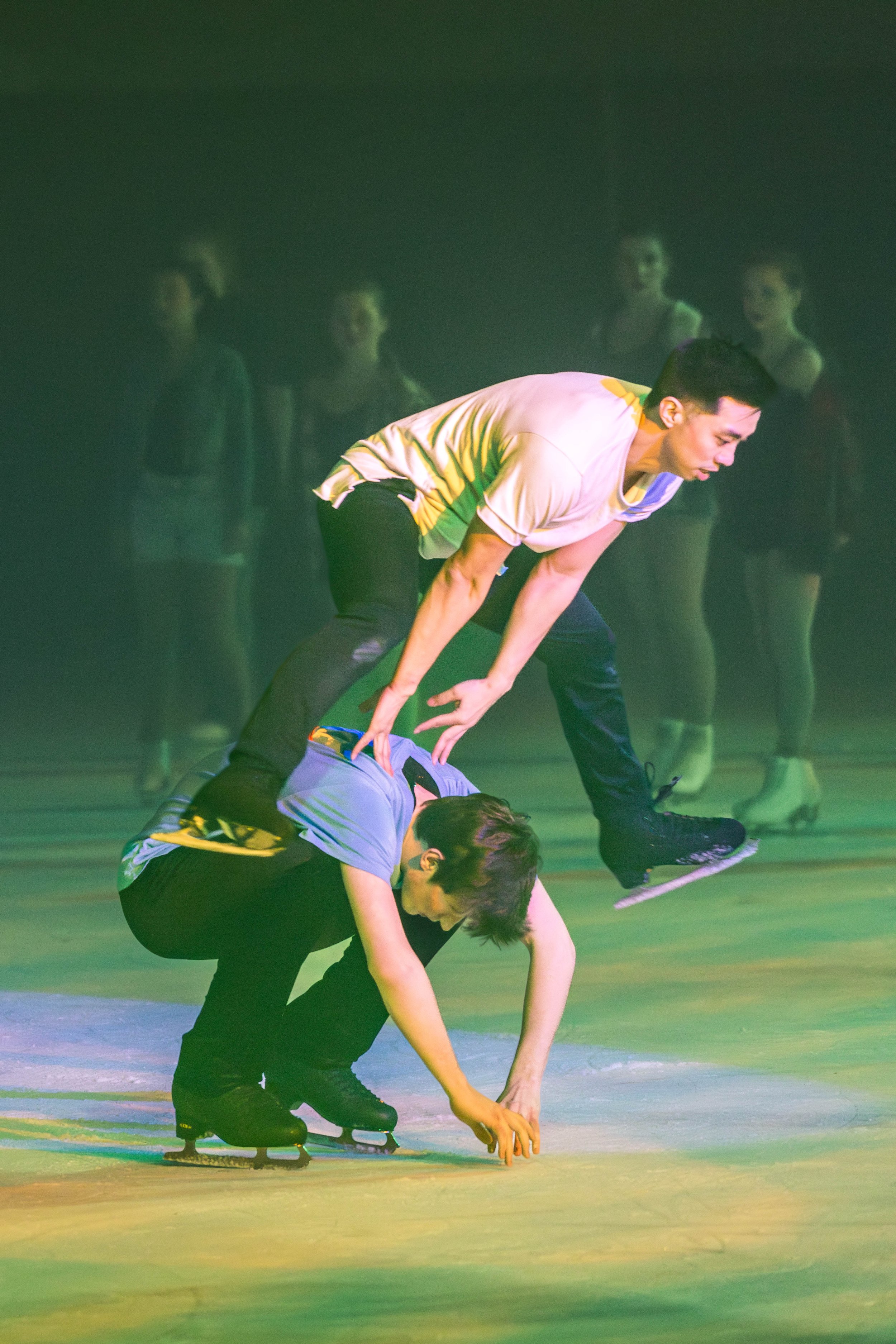 Two young men are figure skating on ice, performing a synchronized move with one bending forward and the other crouching, while four young women in the background observe.