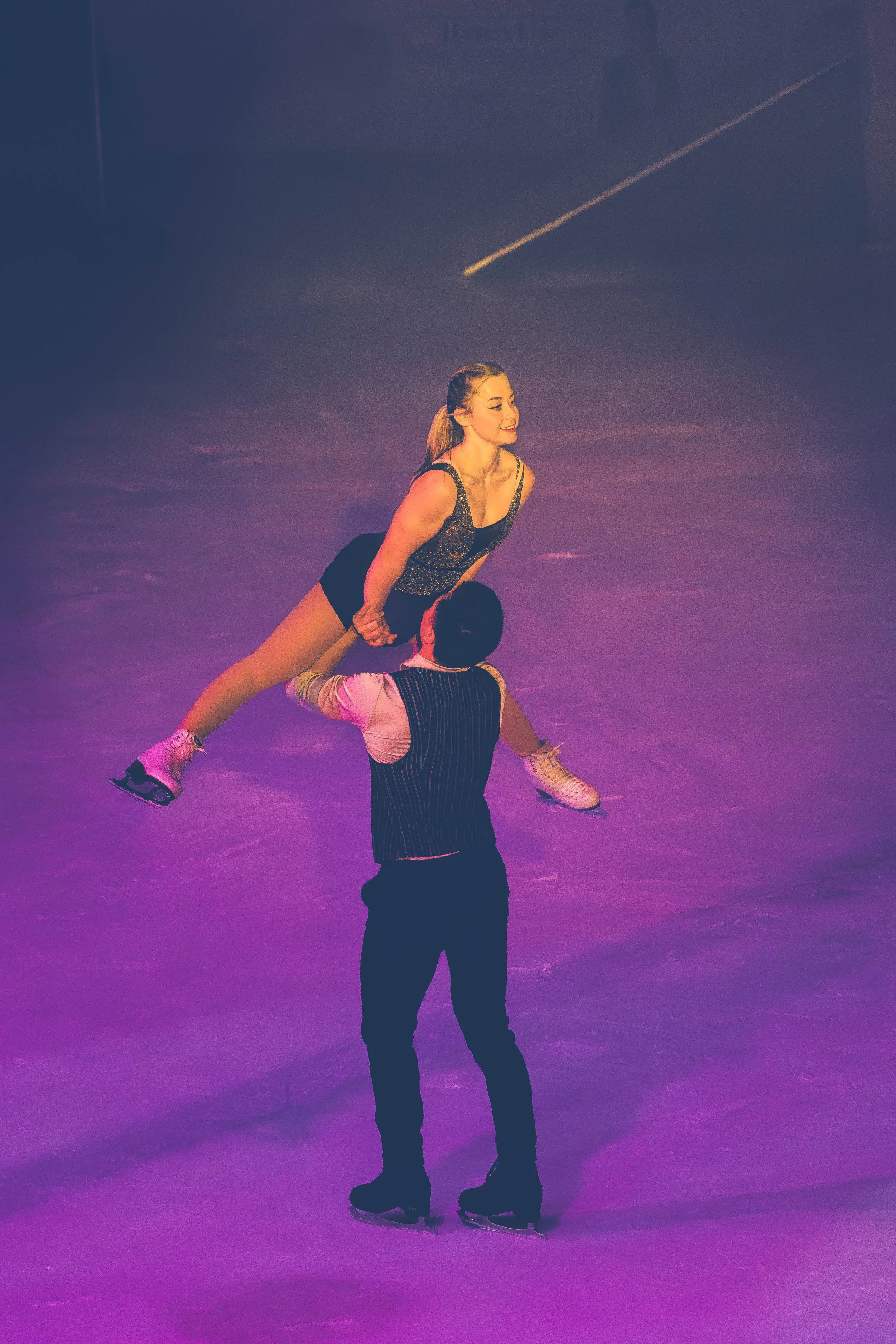 A female figure skater is being lifted by her male partner on the ice rink. The woman appears happy. The scene is illuminated with purple lighting.