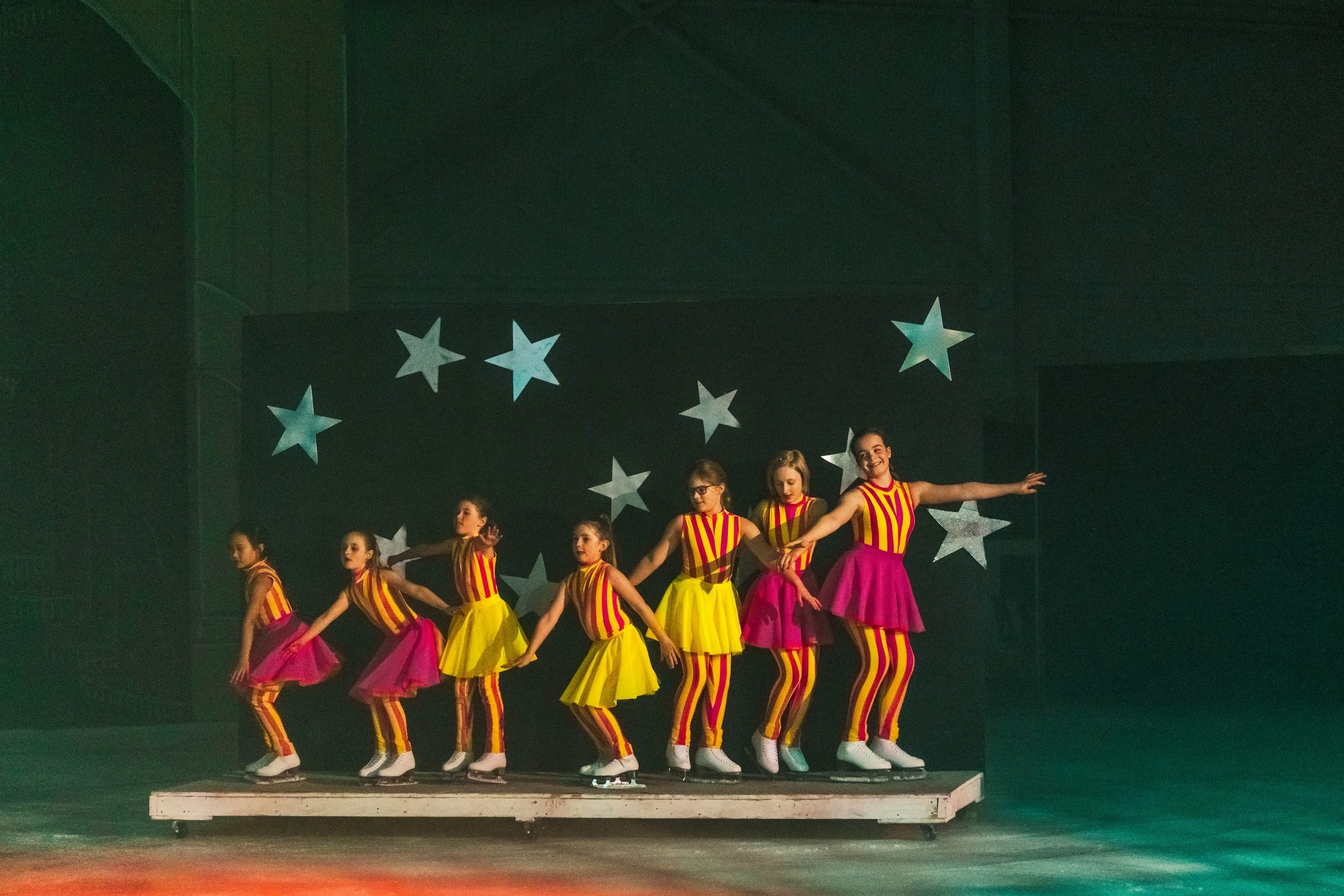 A group of young girls performing a dance on stage in colorful striped costumes with stars in the background.