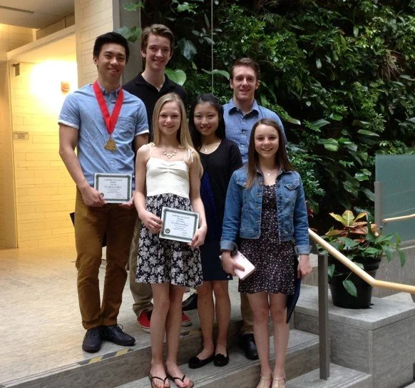 Six young people posing together indoors, with two holding certificates and one wearing a medal, standing in front of green plants.