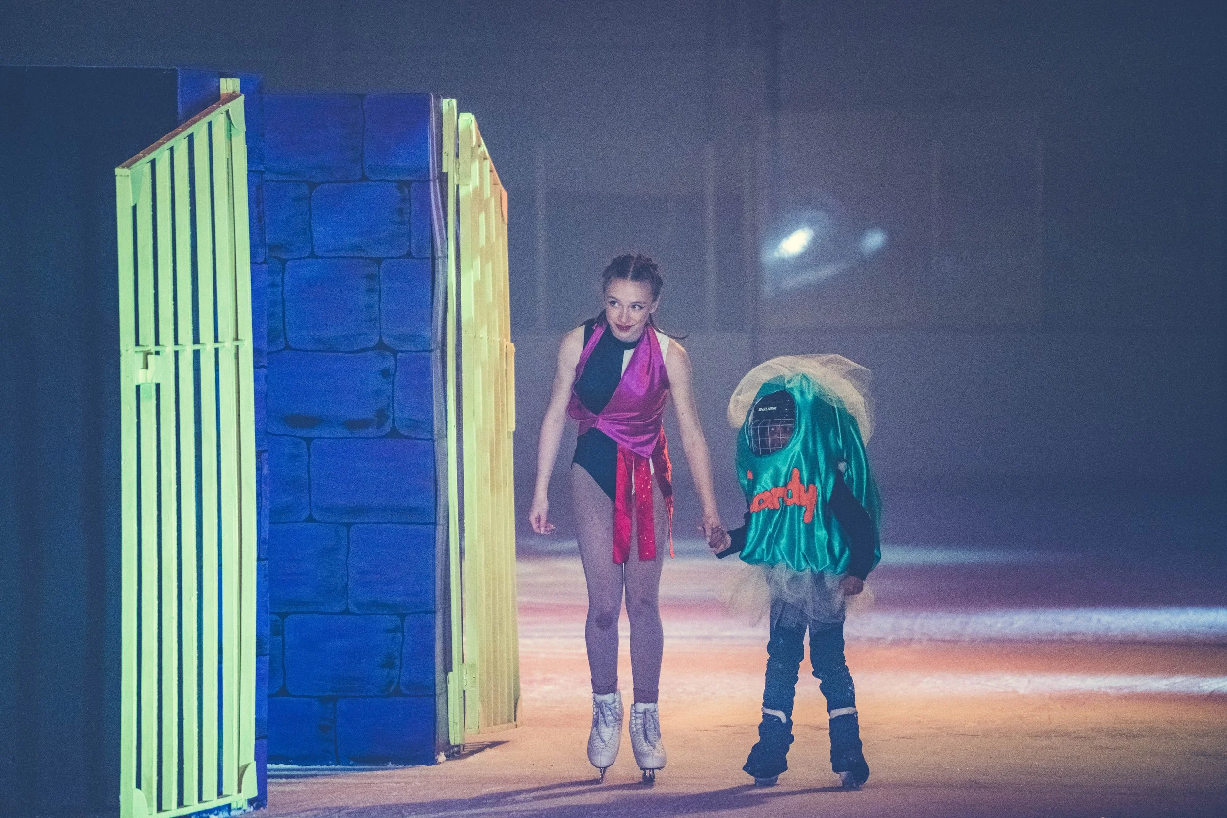 A woman on roller skates holding the hand of a child in a skating rink. The woman is wearing a sleeveless top and shorts, and the child is in a colorful jacket and jeans. There is a brightly colored wall on the left side of the image, and the backgro
