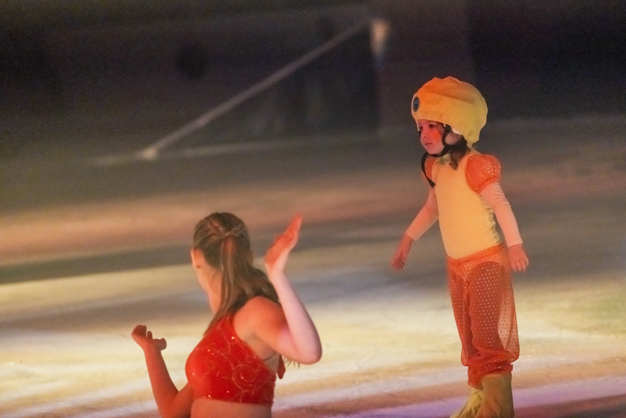 Children in colorful costumes ice skating, one girl is dressed like a duck with a yellow headpiece and orange outfit, another girl is in a red dress, on an indoor ice skating rink.