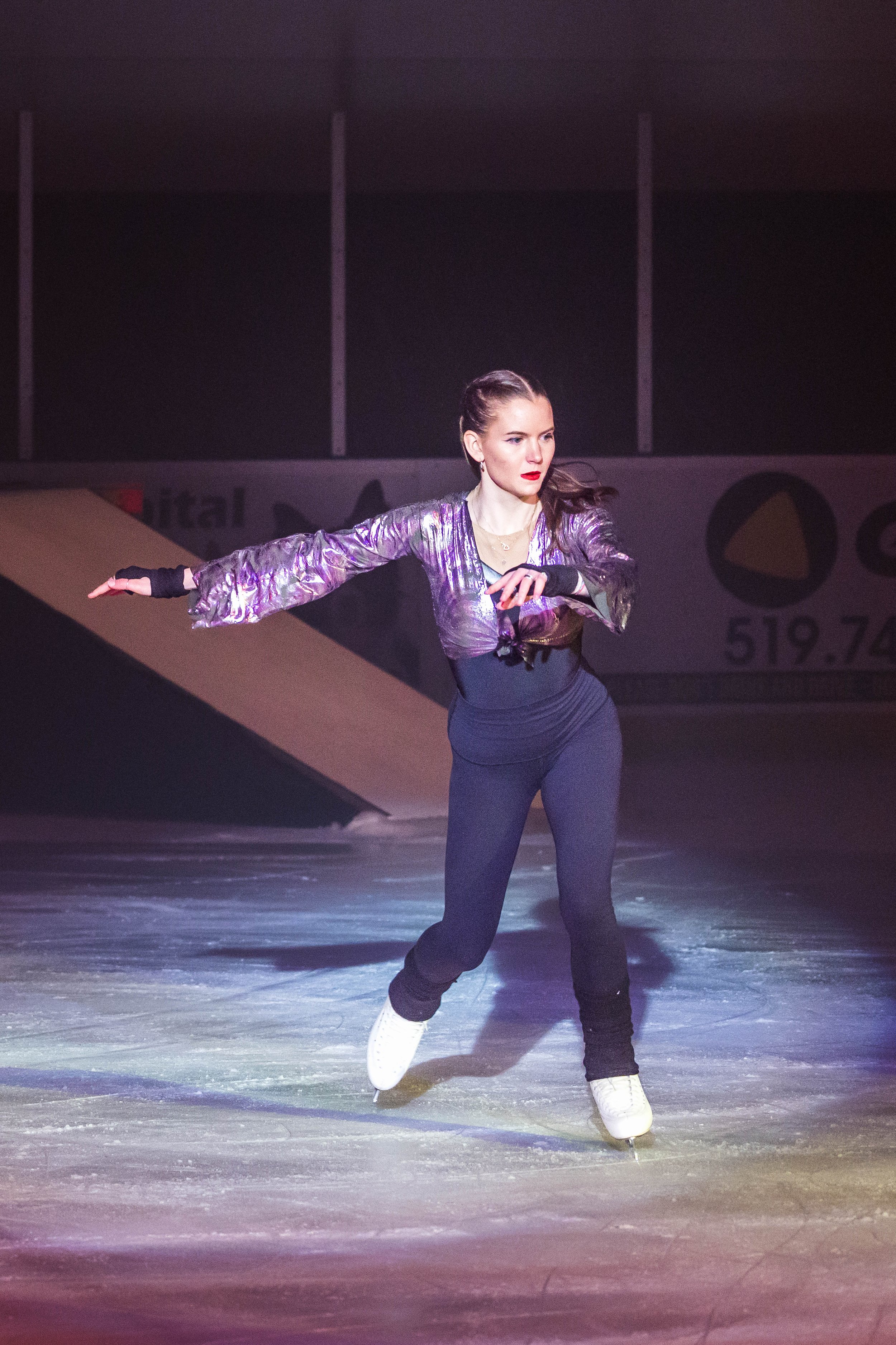 A female figure skater performing on ice, wearing a purple and black costume, with one arm extended and one arm bent, and white ice skates, under bright lighting.