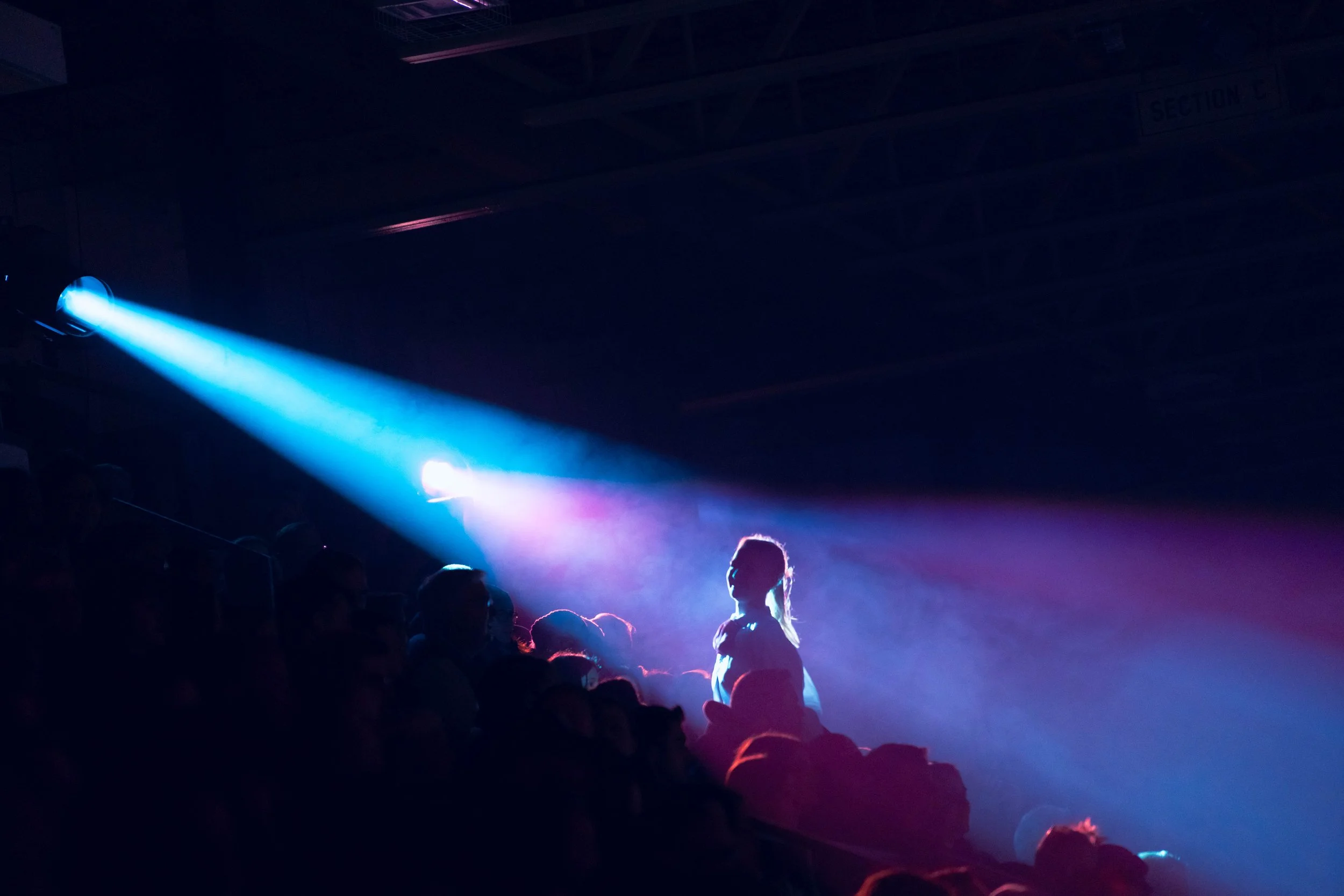 A dark concert or event venue with colorful stage lighting and silhouettes of people in the audience, including a person with glasses standing out in front.